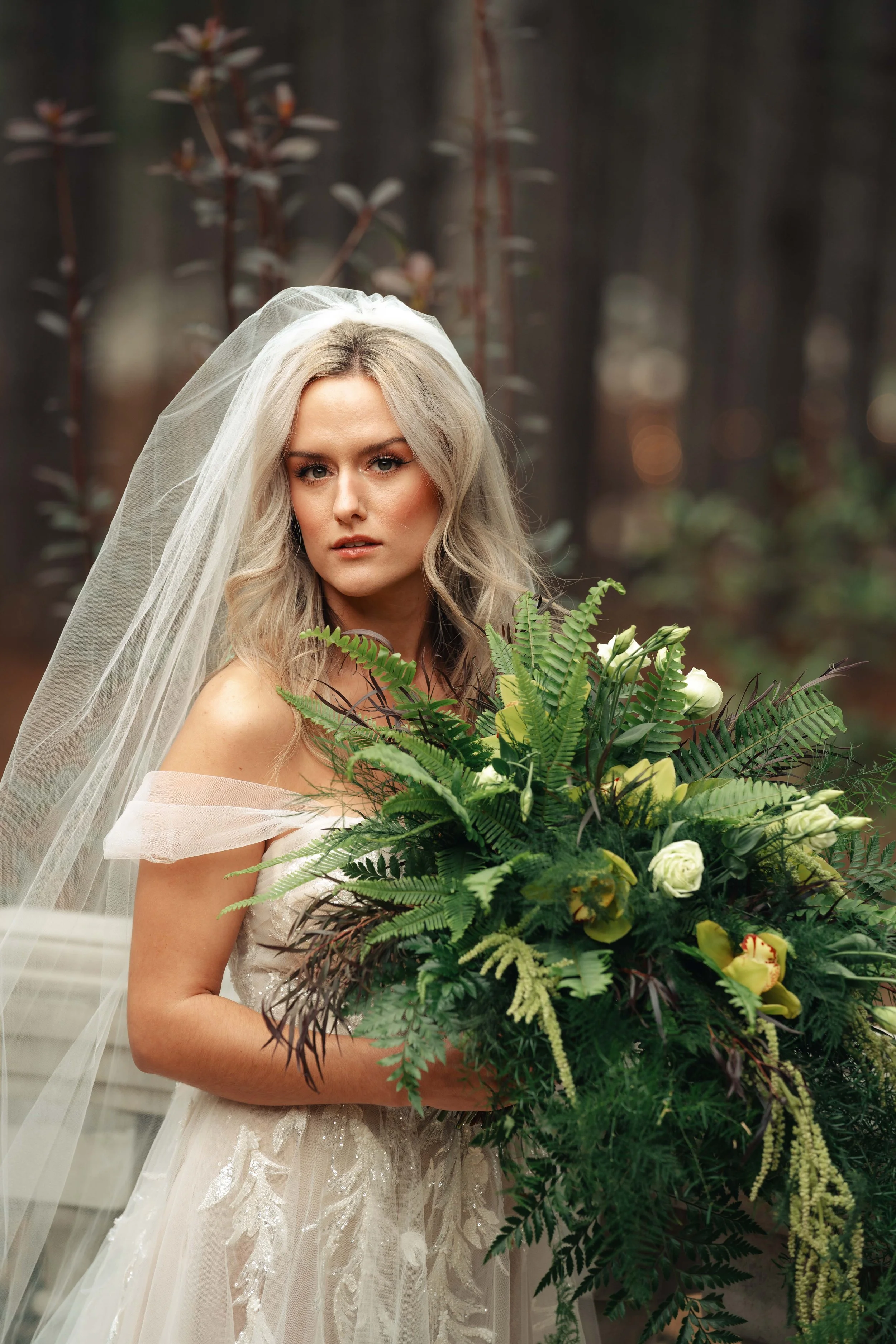 Film camera shot of  bride with blonde wavy hair, wearing a white wedding gown and veil, holding a large bouquet of greenery and white flowers, standing outdoors in forest