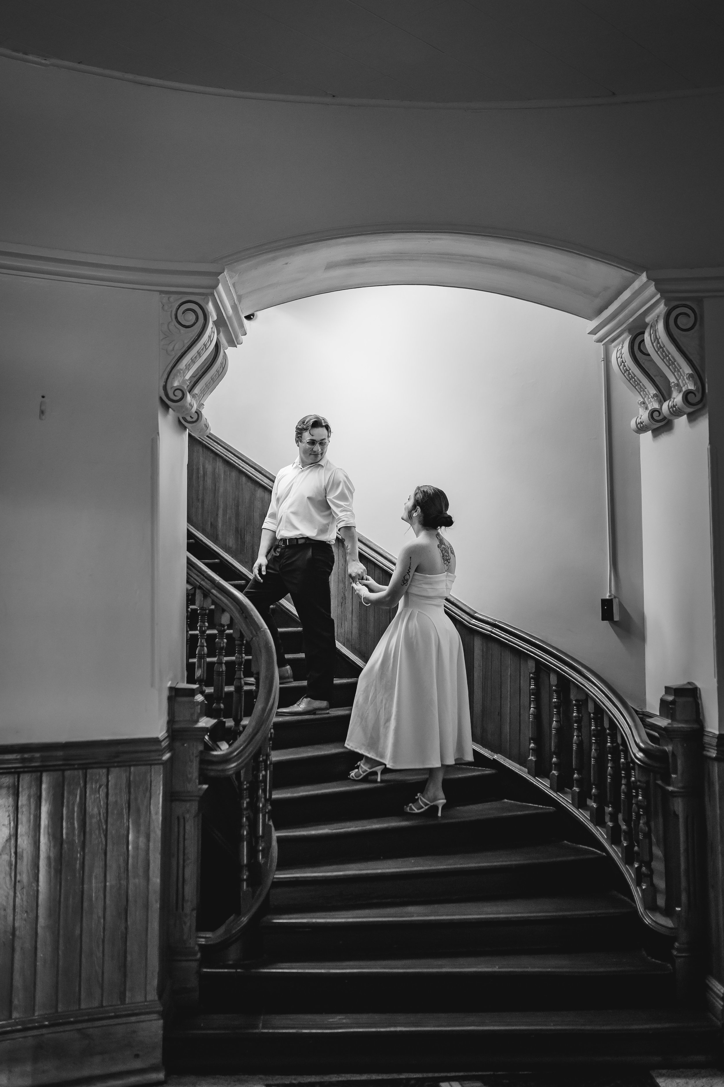 Artistic couple's elopement on wooden staircase, holding hands and looking at each other, dressed in formal attire, with the woman wearing a dress and heels, in an elegant interior setting.