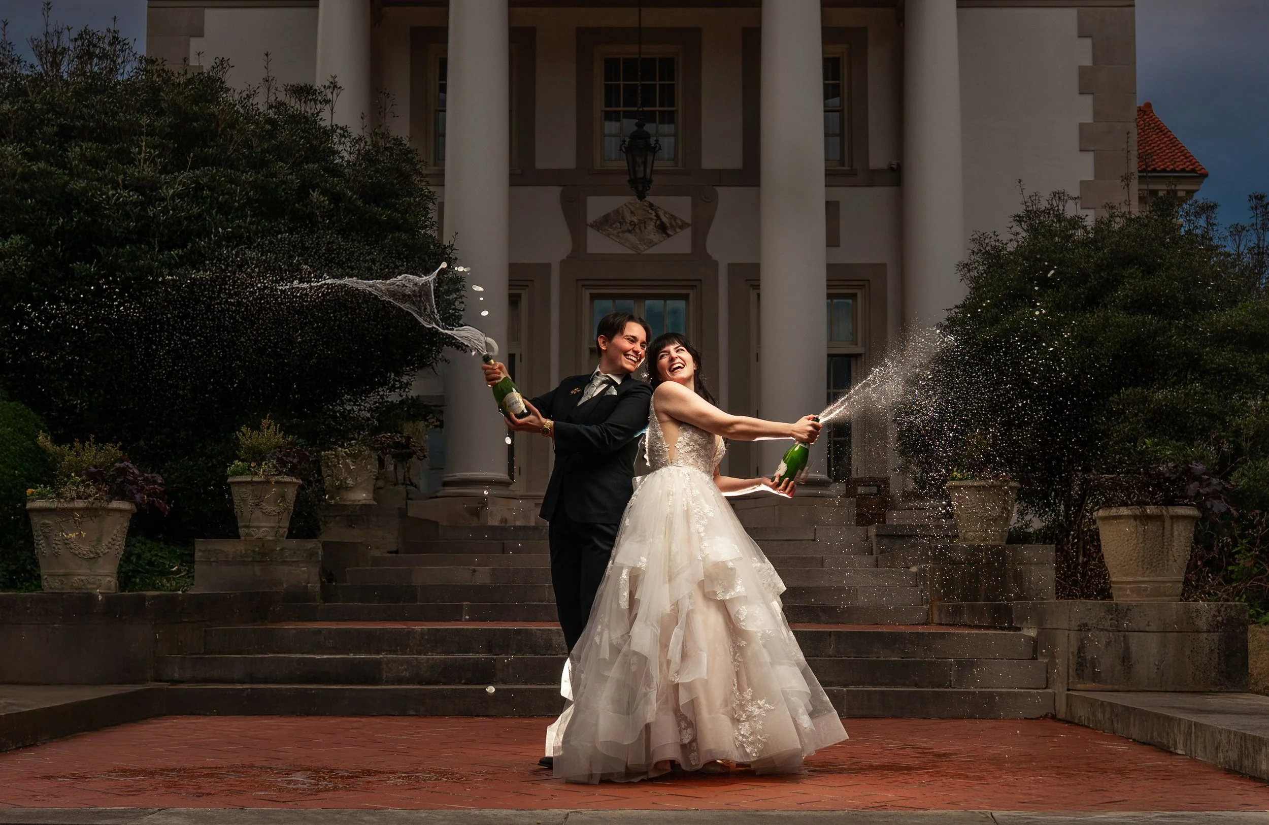 A newlywed couple in wedding attire celebrating with champagne outside a grand building, with both couples spraying champagne into the air, same sex marriage, LGBTQ friendly wedding photographer