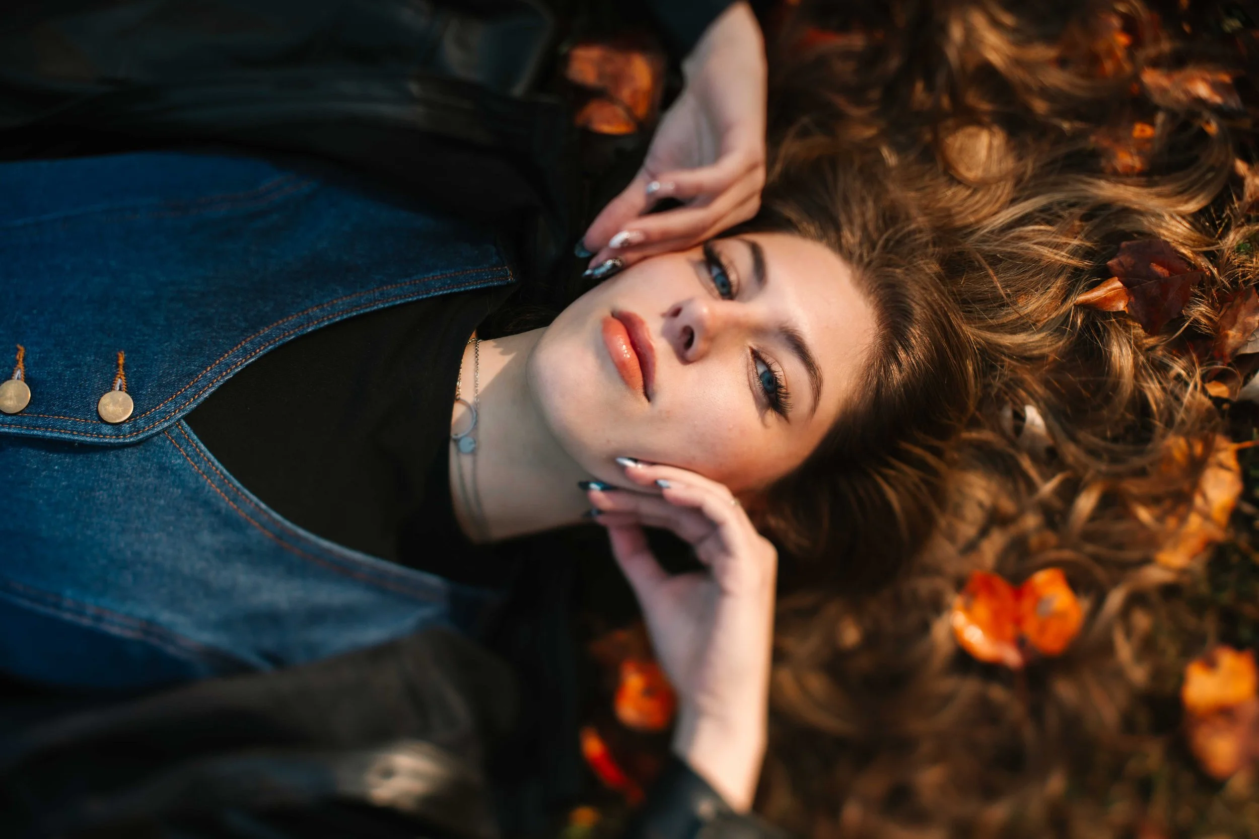 Woman with long, wavy brown hair lying on fallen autumn leaves, wearing a black top and denim vest, looking at the camera.