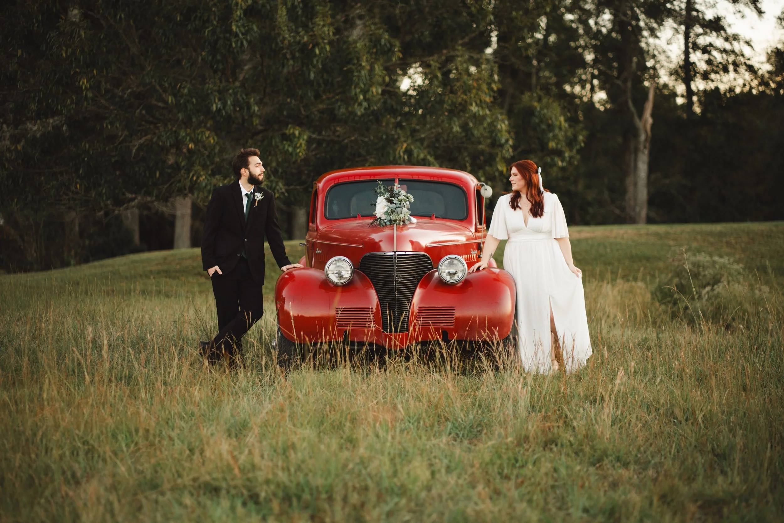 A bride and groom standing beside a vintage red car with a floral arrangement on the hood, in an outdoor grassy field with trees in the background, moody vintage wedding photography