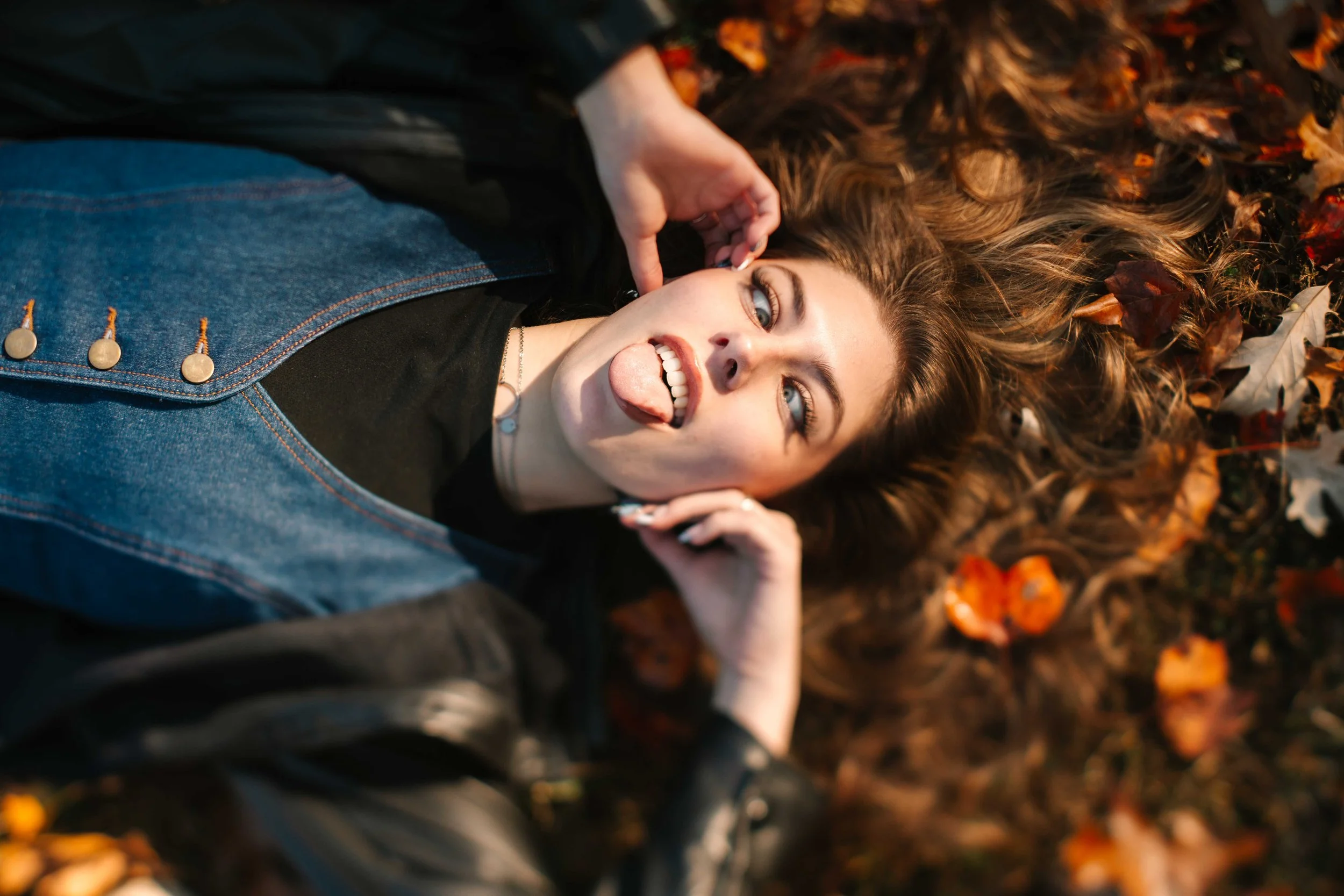 Woman lying on autumn leaves, sticking out her tongue, with long wavy hair spread around her, wearing a black leather jacket, denim shirt, and silver jewelry.