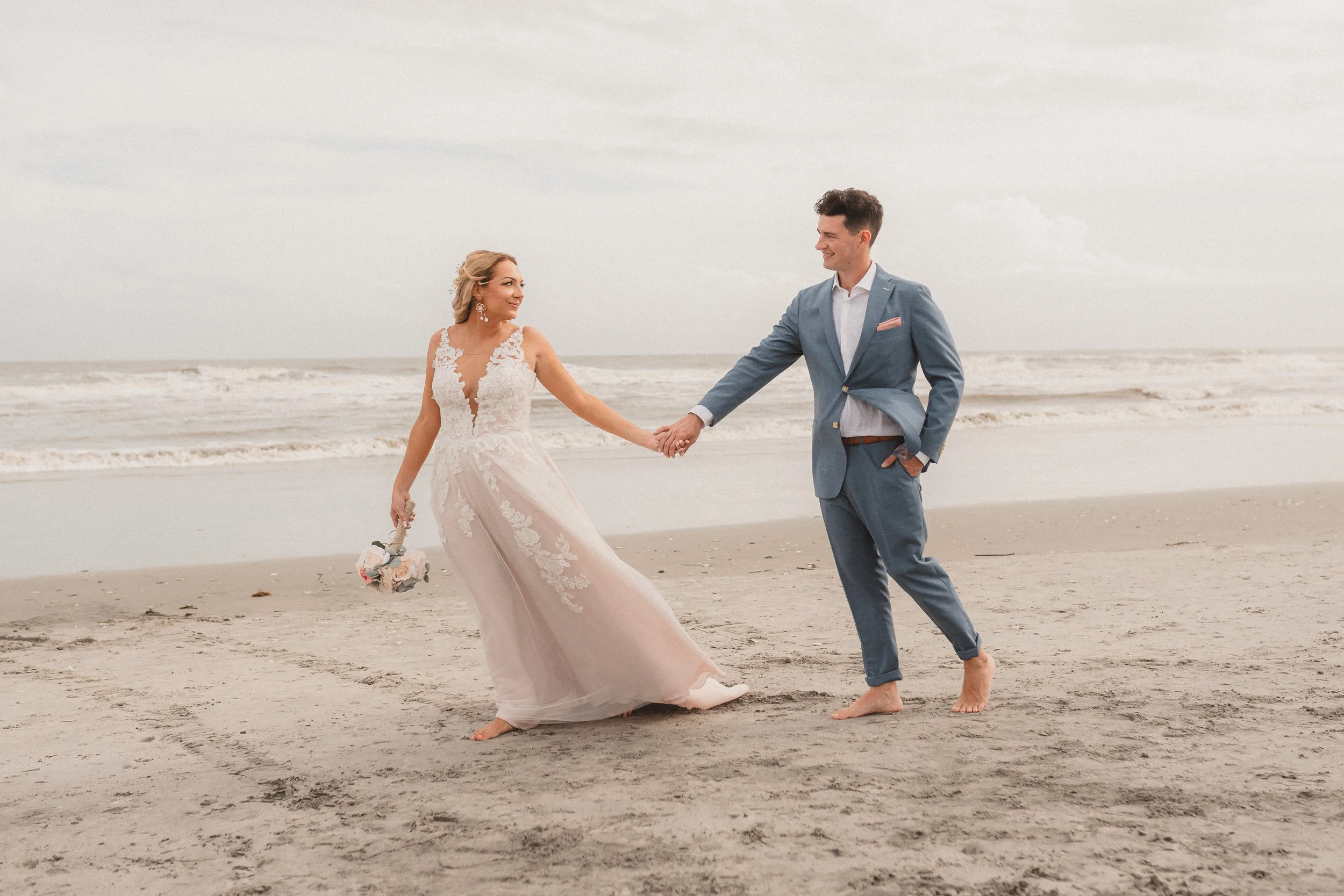 A bride and groom holding hands and smiling on a sandy beach with the ocean in the background, both barefoot, candid beach wedding photo of couple running across the shoreline