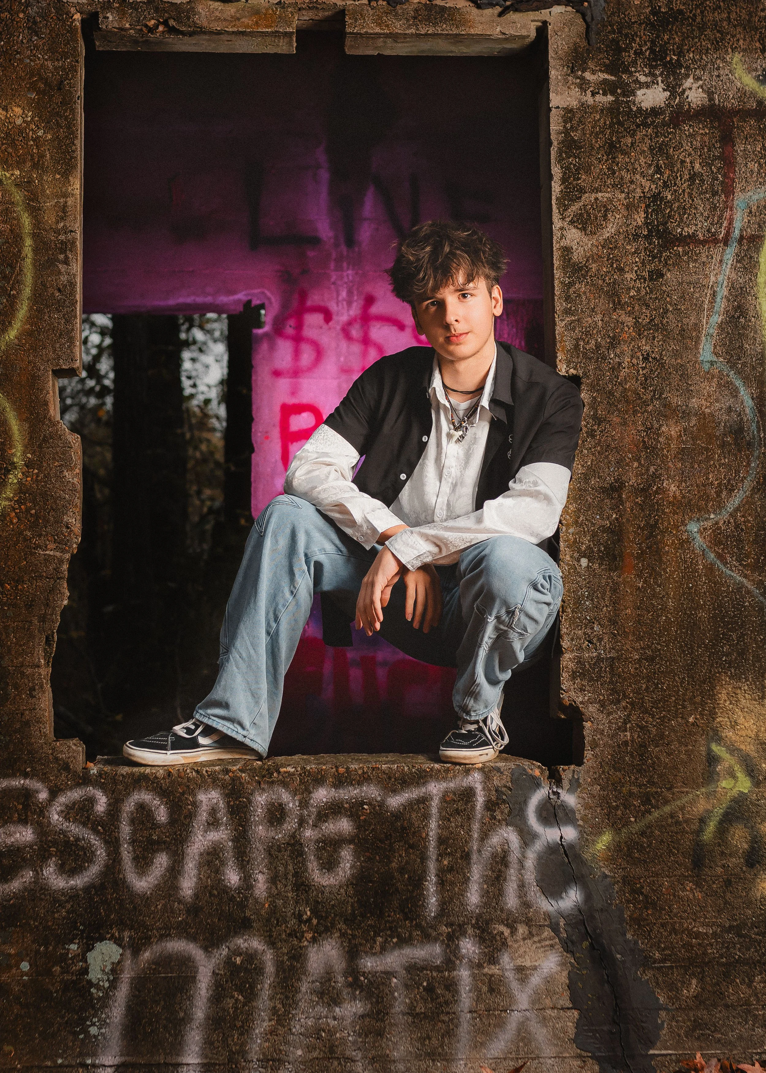 A young man with curly hair, dressed in a white shirt with a black jacket and light-wash jeans, is squatting in a graffiti-covered, abandoned building frame with a pink spray-painted background.