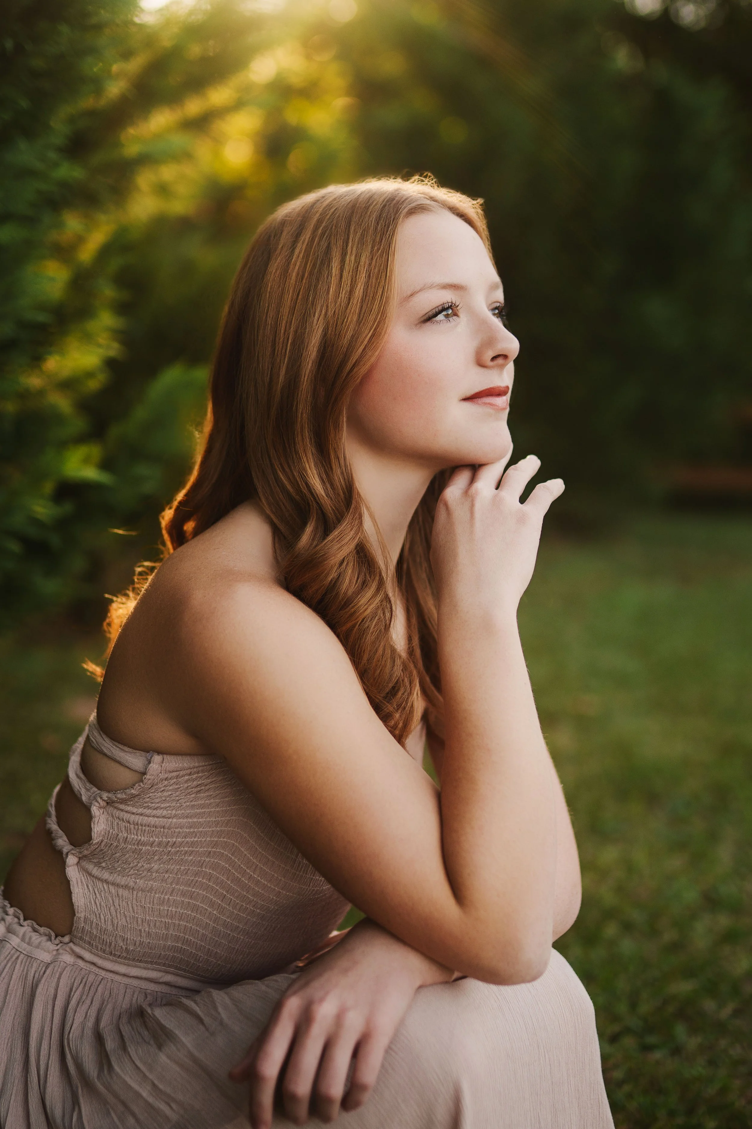 A woman with red hair sitting outdoors in a green, wooded area during sunset, looking thoughtfully to the side.