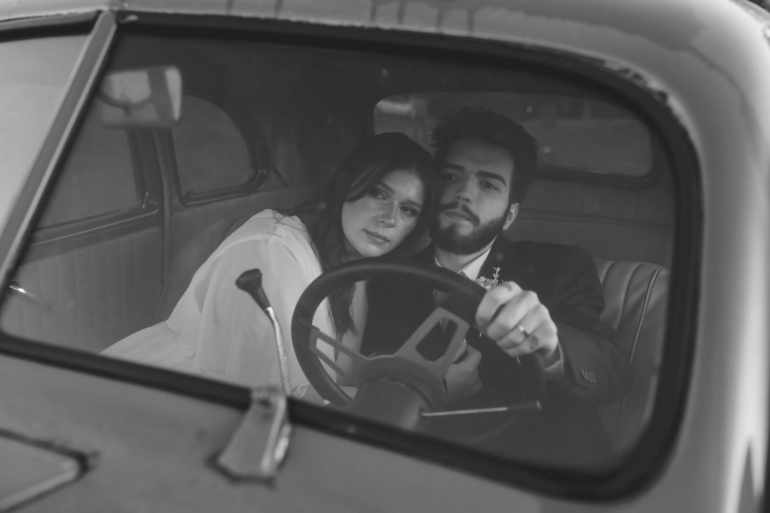 Documentary style wedding photography, Black and white photo of a couple sitting inside a vintage car, looking through the front windshield. 