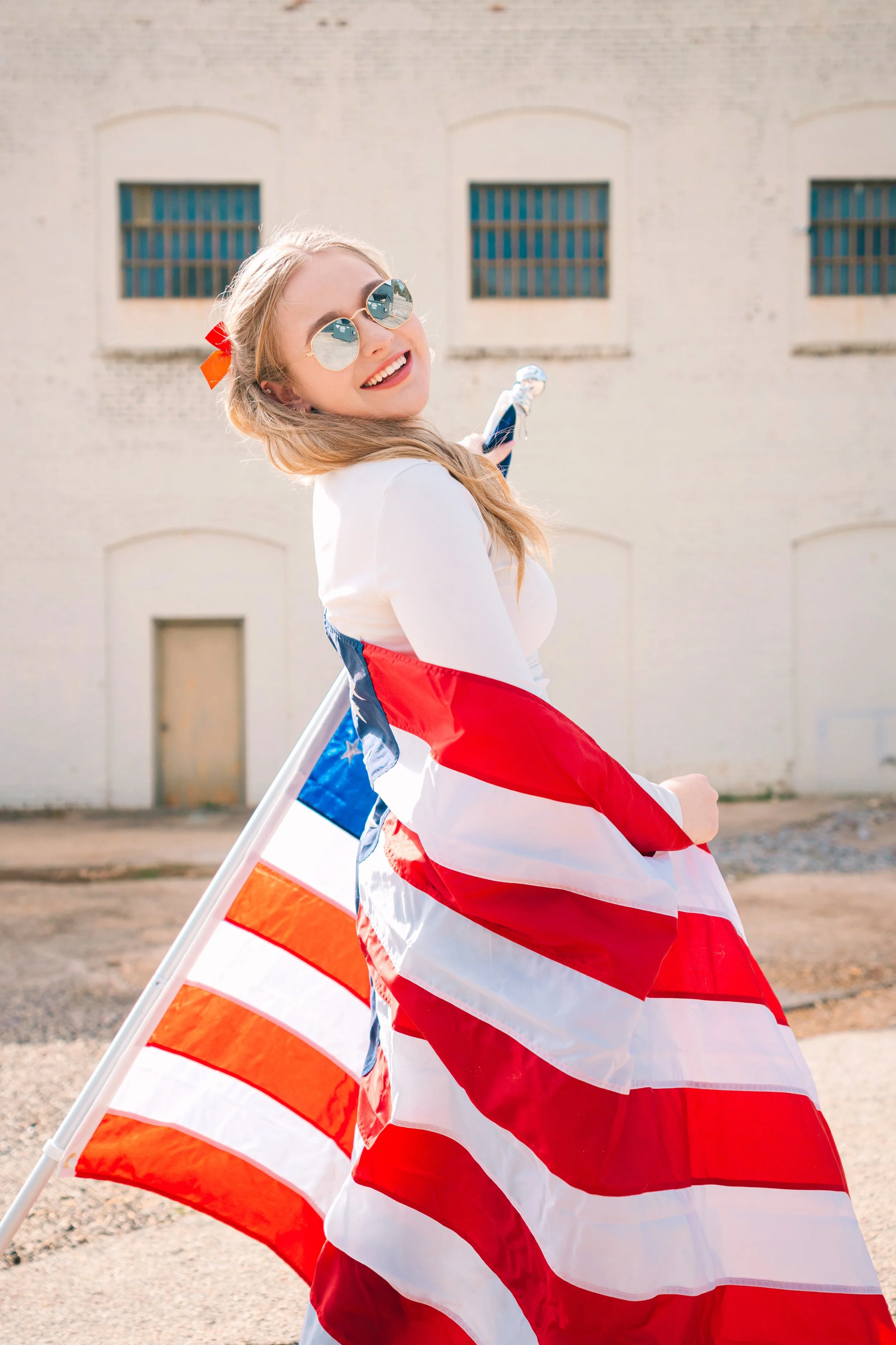 Fourth of July and Memorial Day Weekend photo session, wearing sunglasses and a white shirt, holding a French flag, smiling and standing outdoors in front of a white building.