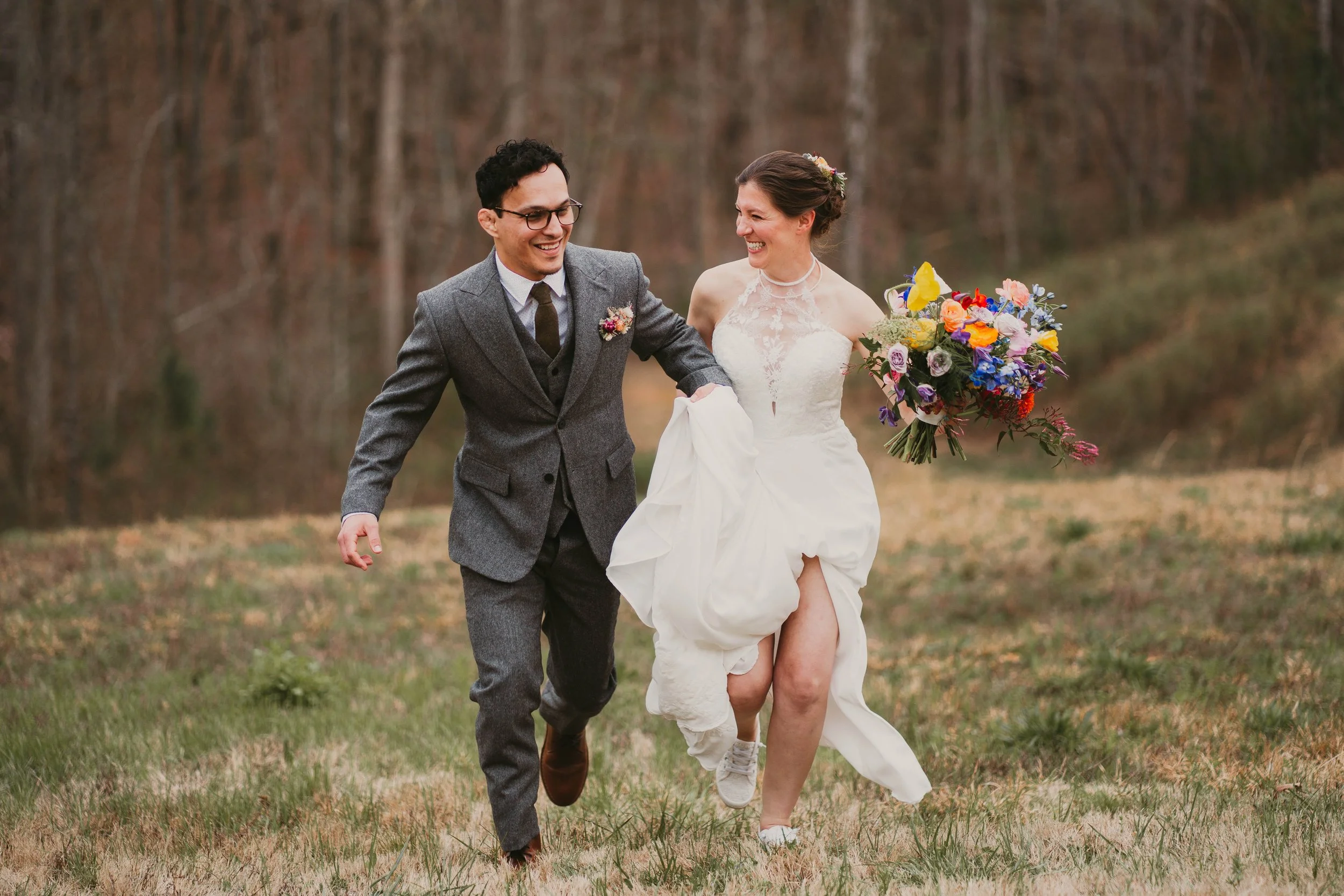 A newlywed couple running through a grassy field, smiling and holding hands, with a colorful bouquet of flowers, during daytime in a wooded area, whimsical cottagecore weddings