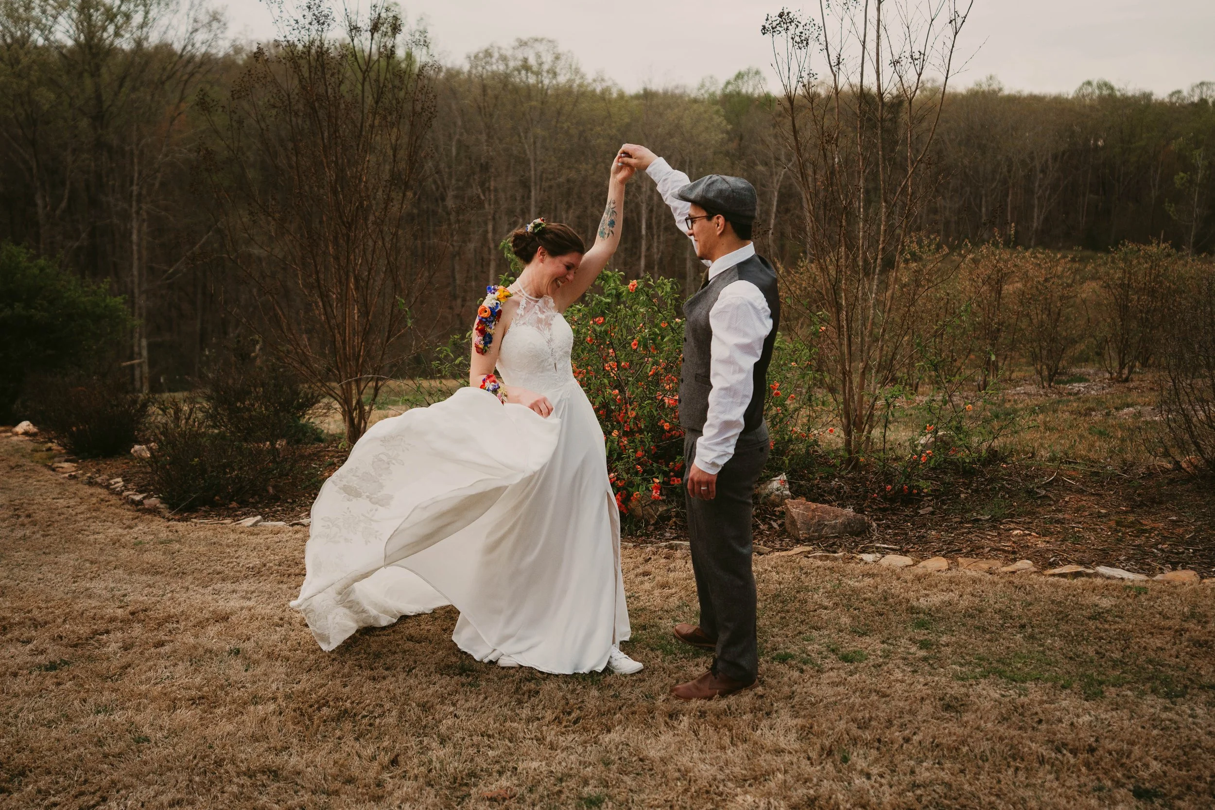A bride and groom dancing outdoors in a natural setting with trees and bushes, one woman in a white wedding dress and the man in a white shirt, gray vest, and gray hat, holding hands and smiling.