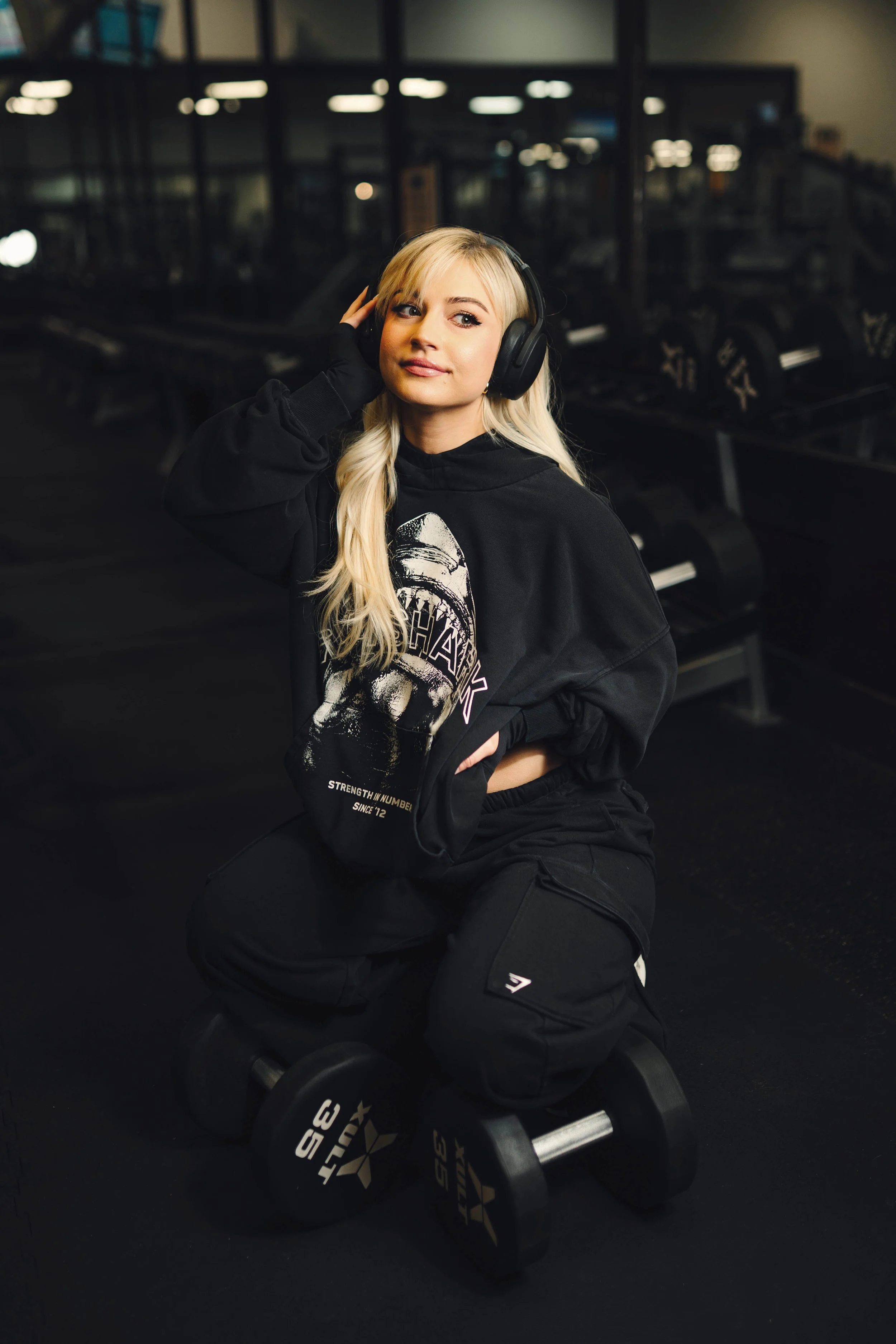 A young woman with long blonde hair kneeling in a gym, wearing a black hoodie listening to music with large over-ear headphones. There are dumbbells on the floor in front of her. Modern fitness photo session, fitness influencer