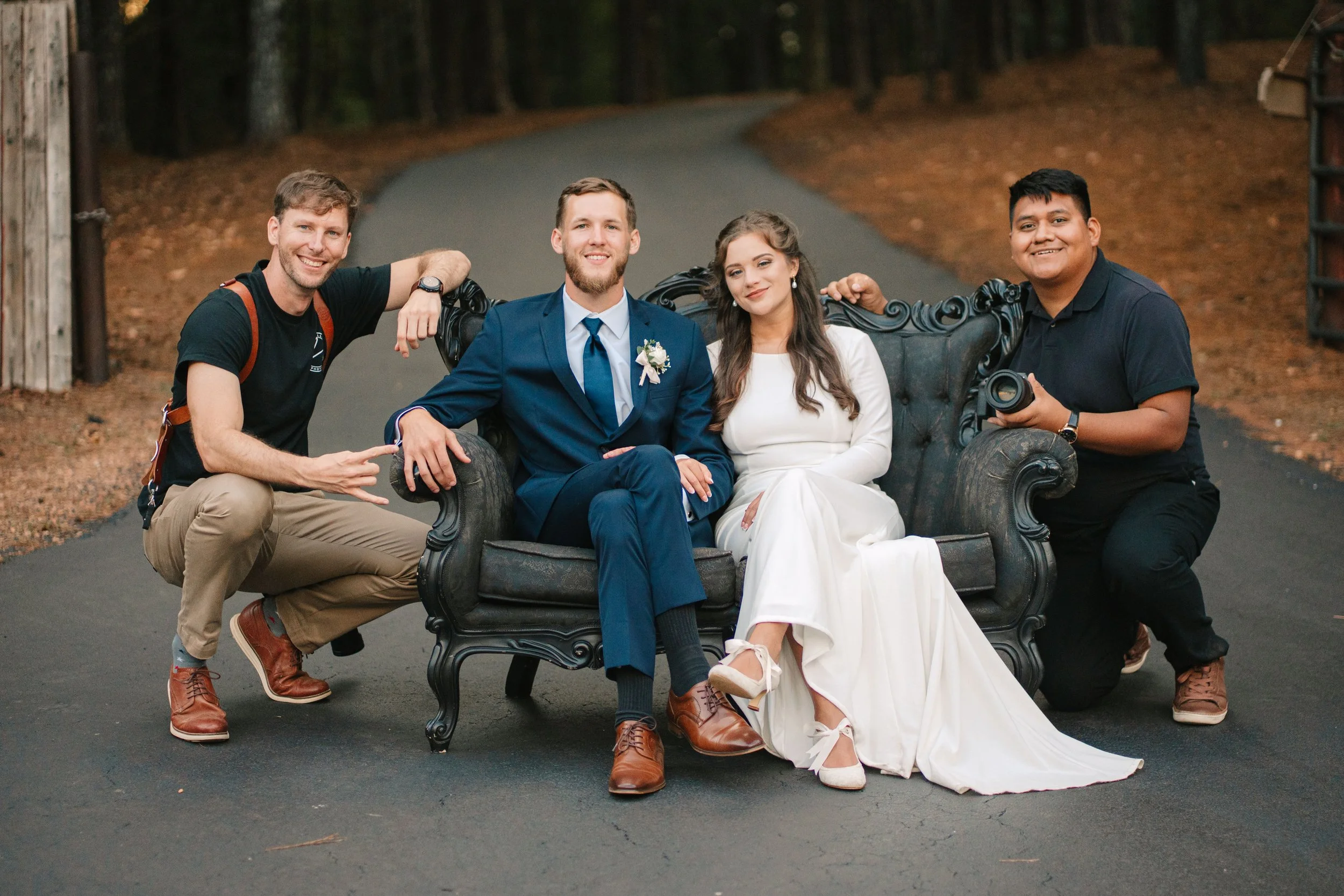 A wedding couple sitting on a vintage black sofa outdoors with photographers kneeling beside them, on a paved road surrounded by trees and autumn leaves.