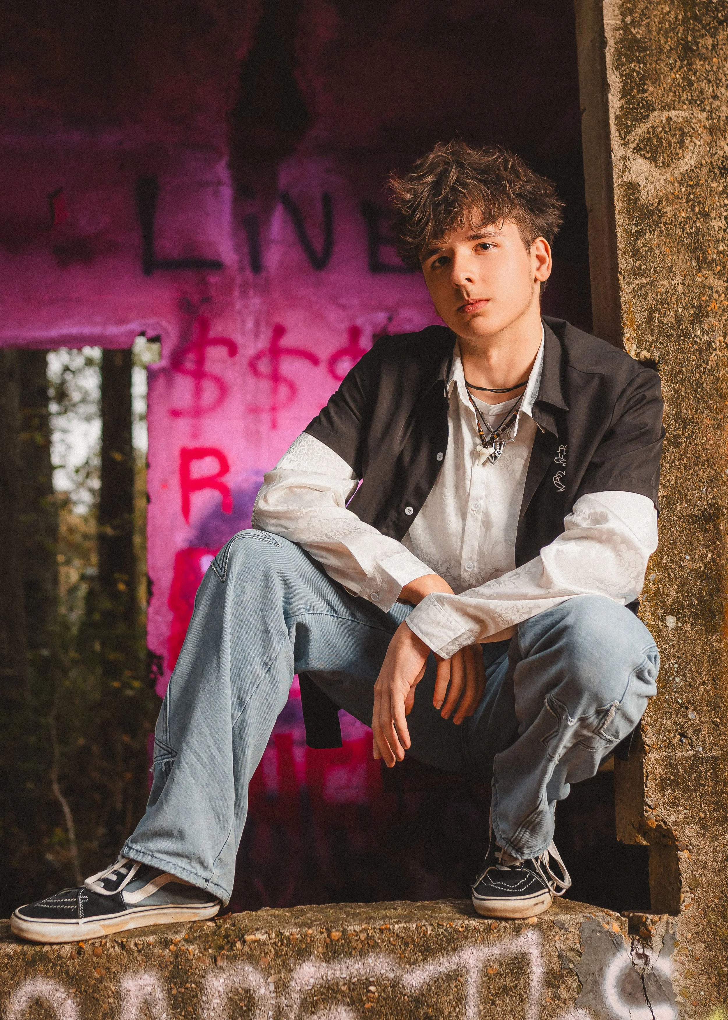 A young man with curly hair dressed in casual clothes, sitting on a ledge in front of a graffiti wall in an urban, outdoor setting.