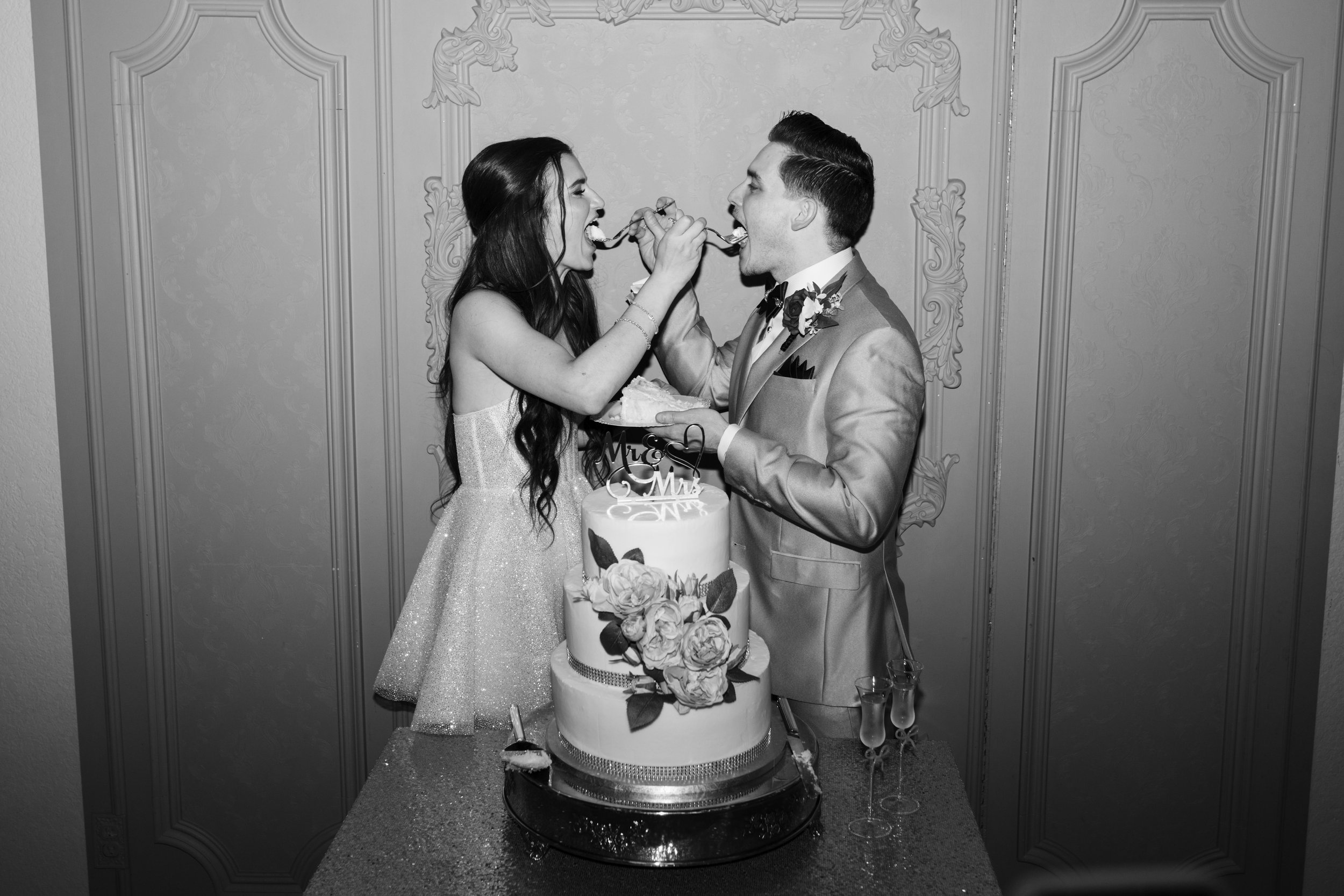 Black and white photo of a newlywed couple feeding each other cake at their wedding reception, standing behind a decorated wedding cake on a table, with ornate wall decor in the background.