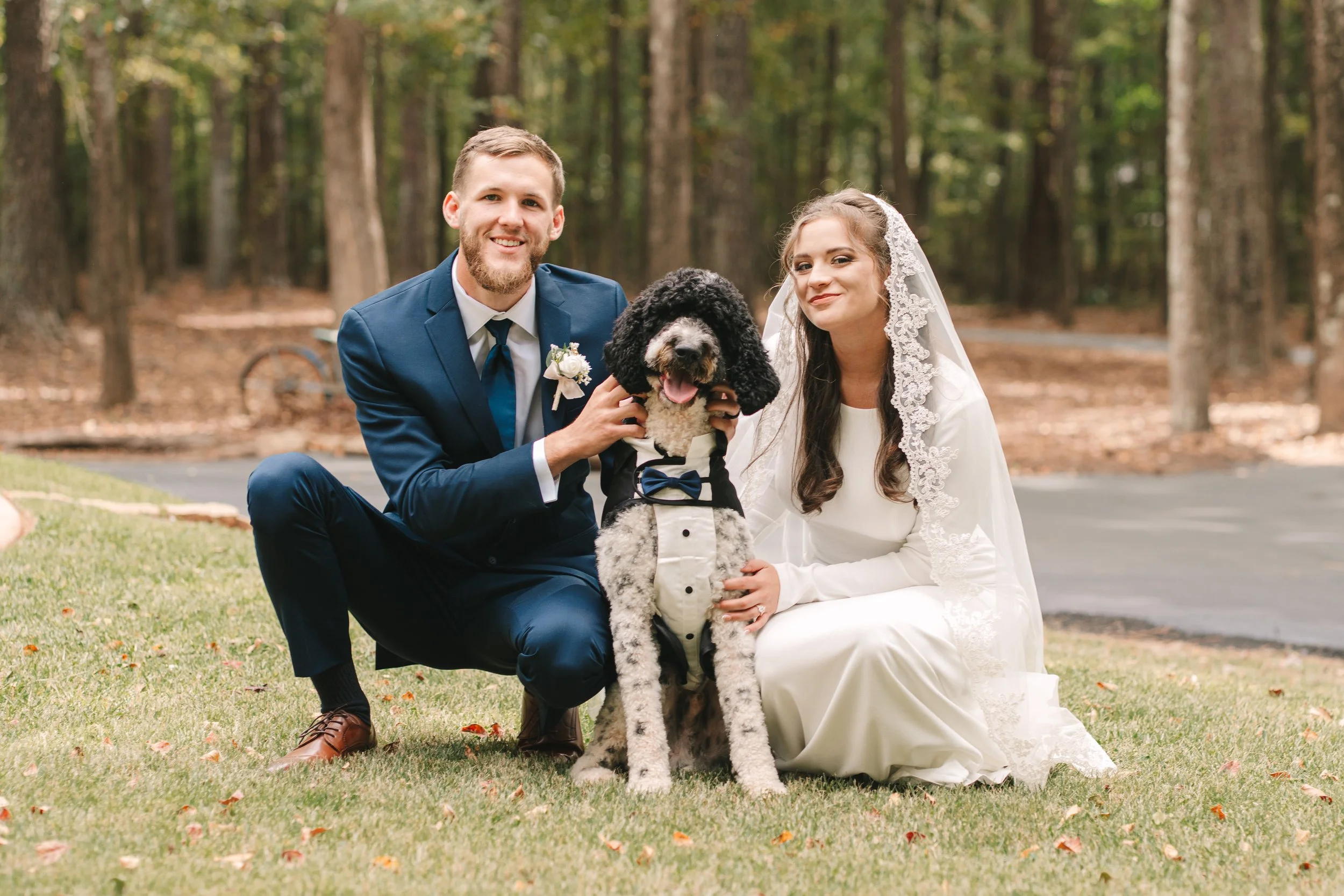 Decatur, GA newlywed couple with a large black and white poodle dressed in a tuxedo, posing outdoors in a wooded area.