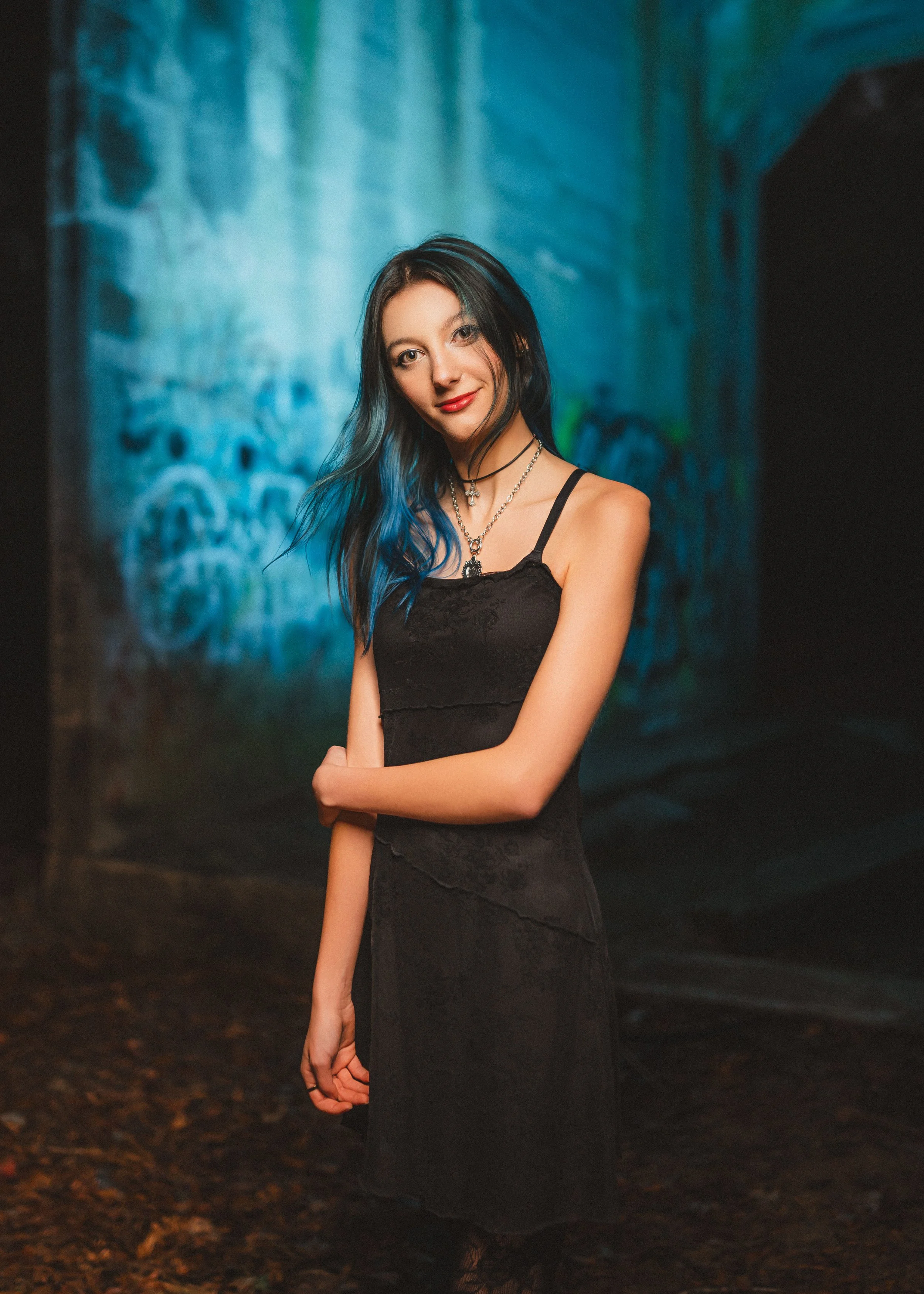 A young woman with black and blue hair in a black dress and layered necklaces standing in front of a graffiti-covered wall in a dark alley. Smoky punk rock vibe