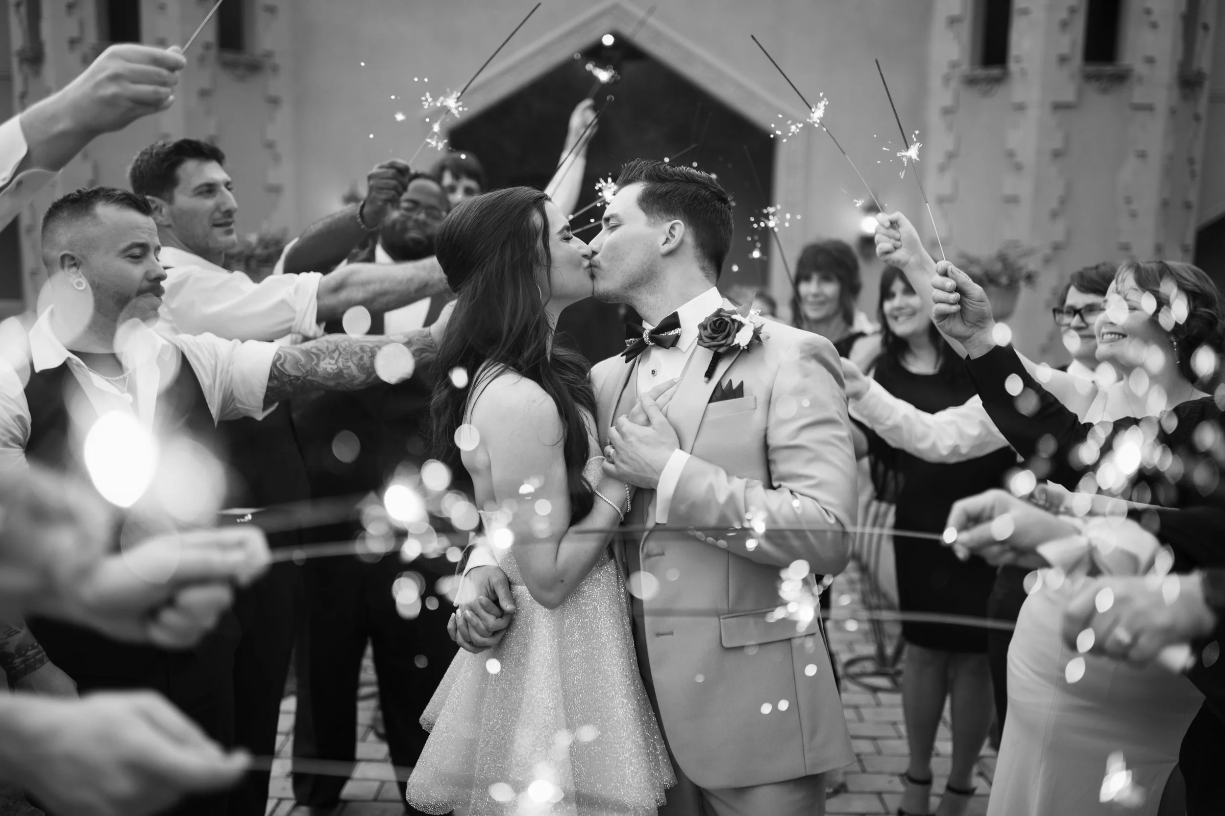 Fairytale wedding venue, couple shares a kiss as friends and family celebrate, holding sparklers around them in a romantic, joyful moment during their wedding reception.