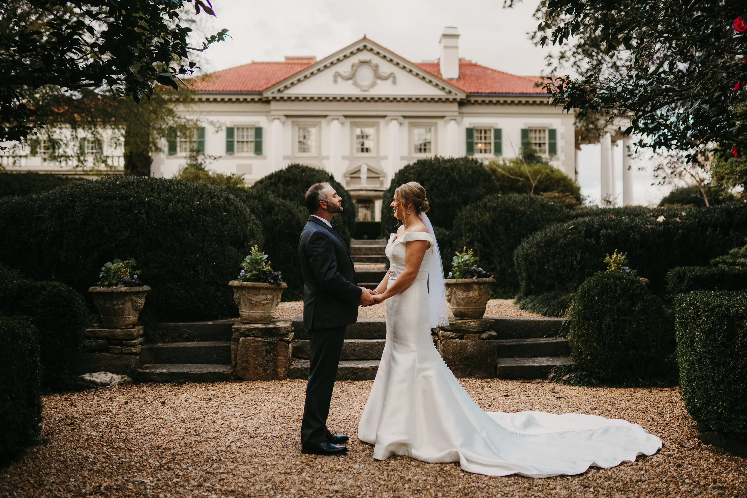 A bride and groom holding hands and looking at each other in front of a large, elegant white house with green shutters and a red-tiled roof, surrounded by shrubbery and potted plants.