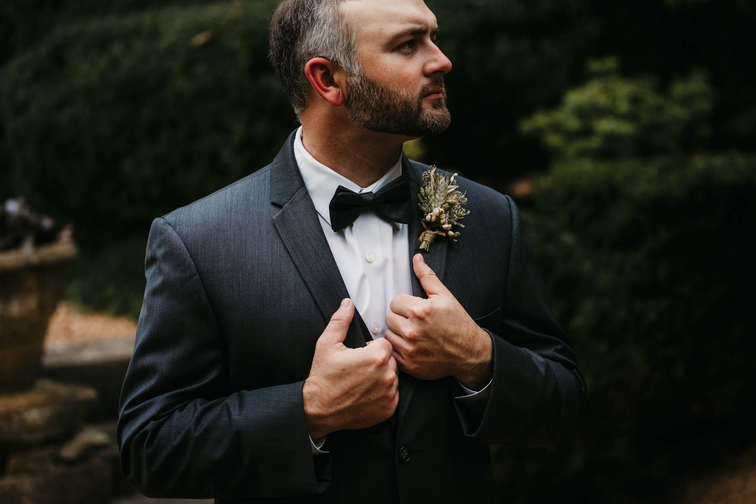 Groom in a tuxedo with a bow tie and boutonniere adjusting his jacket outdoors, moody wedding detail shot