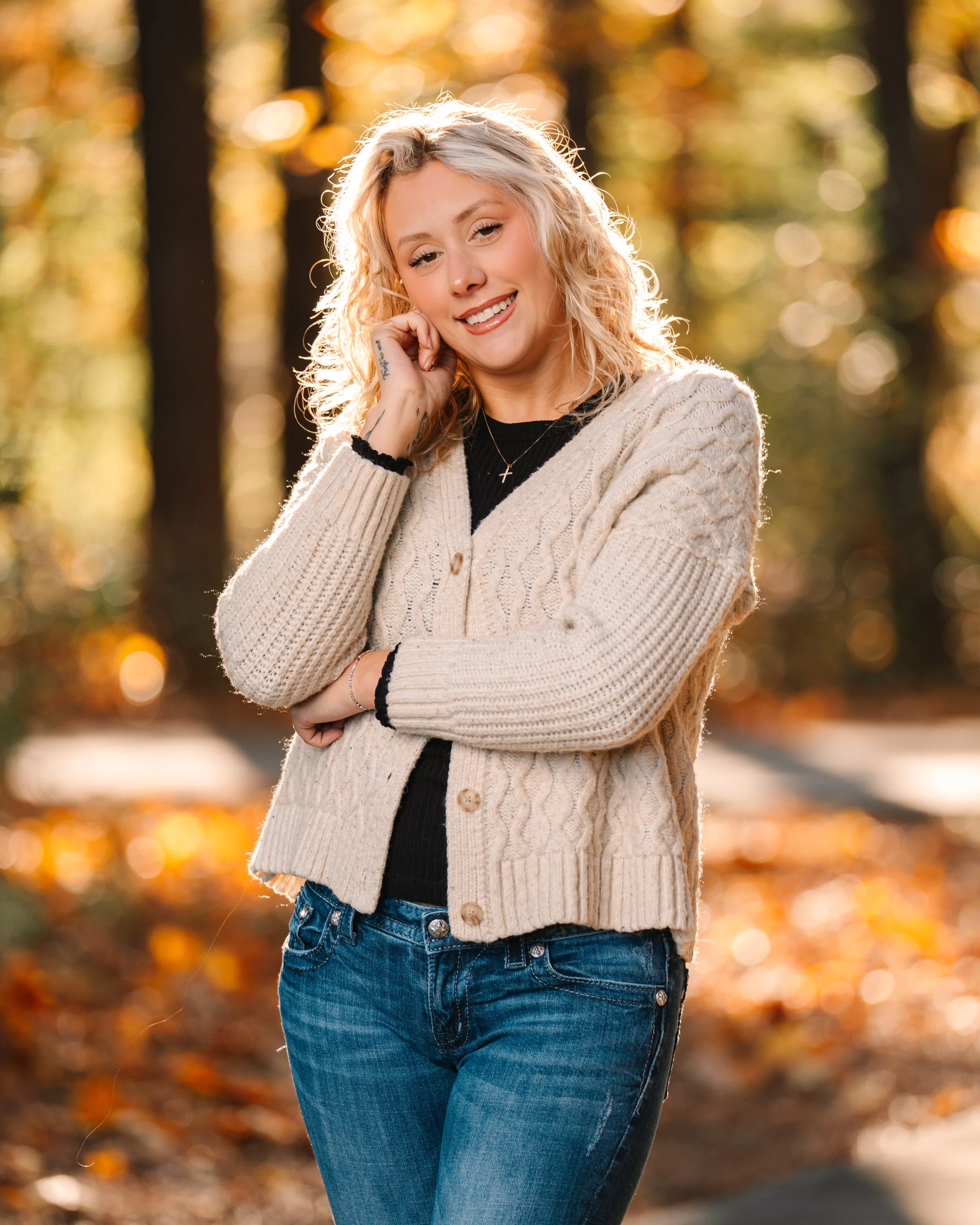 A young woman with blonde curly hair smiling outdoors in autumn, wearing a beige knit cardigan and blue jeans, with sunlight filtering through trees in the background.