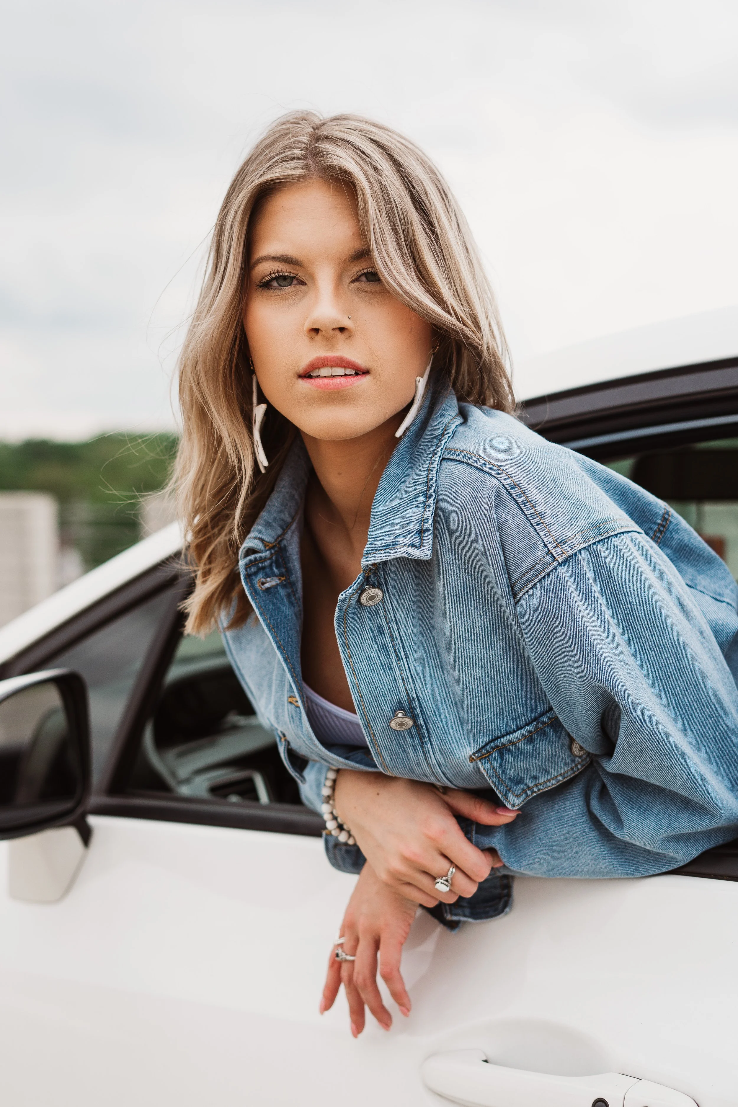 Trendy Teen with wavy blonde hair leaning out of a white car window, wearing a denim jacket, earrings, and rings.