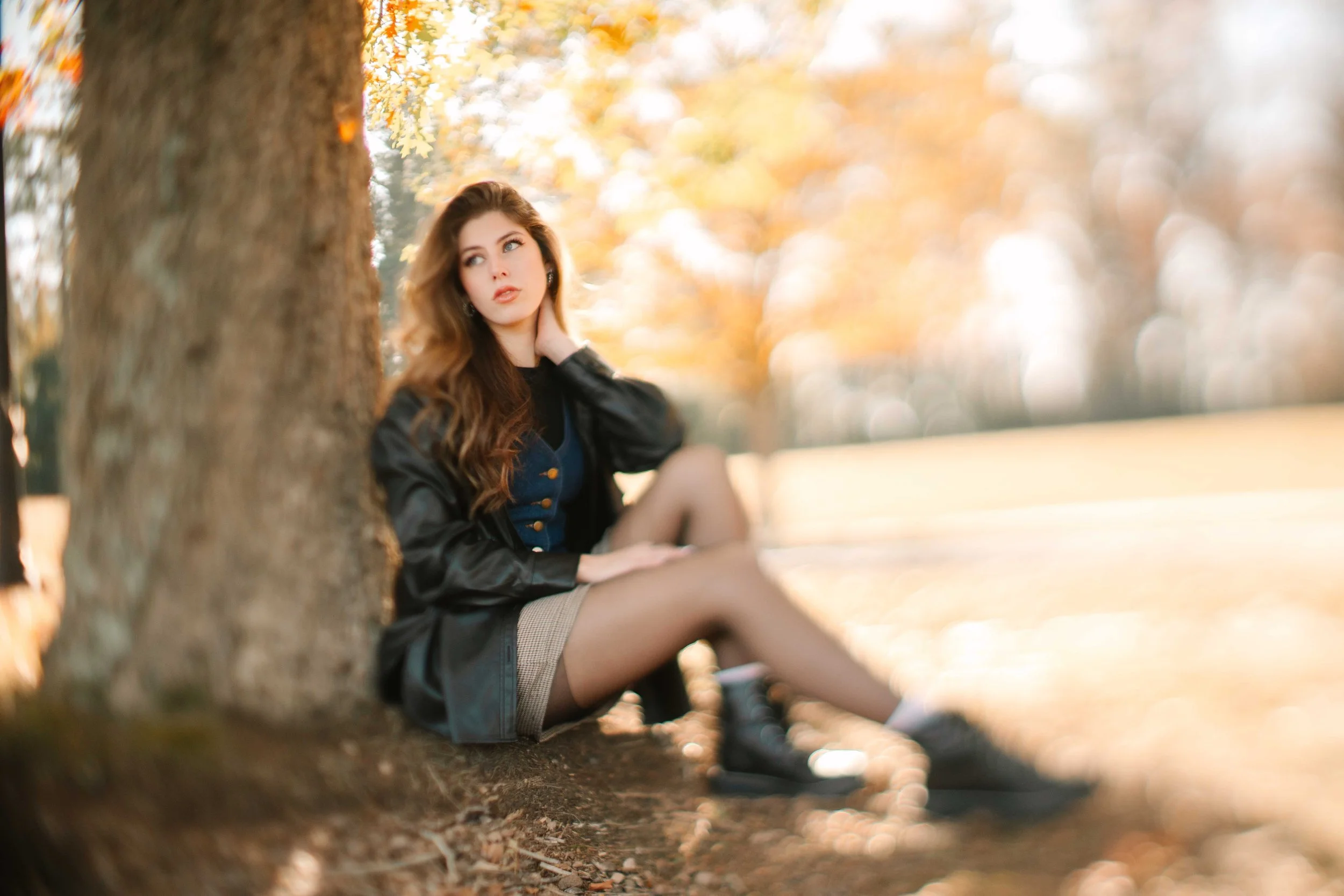 Fall mini session with young woman with long, wavy brown hair sitting on the ground next to a tree in a park during autumn. She is wearing a black jacket, blue top, plaid skirt, and black combat boots