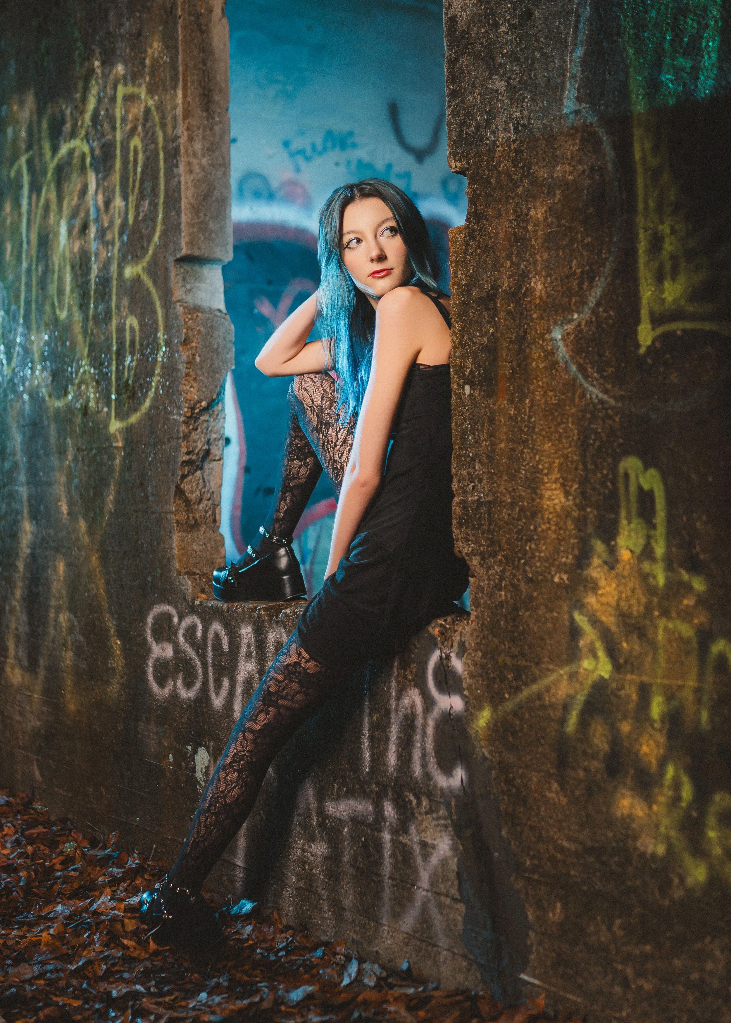 A young woman with blue hair sitting on a concrete wall with graffiti, surrounded by graffiti-covered walls and fallen leaves on the ground. Edgy and grunge style senior portrait