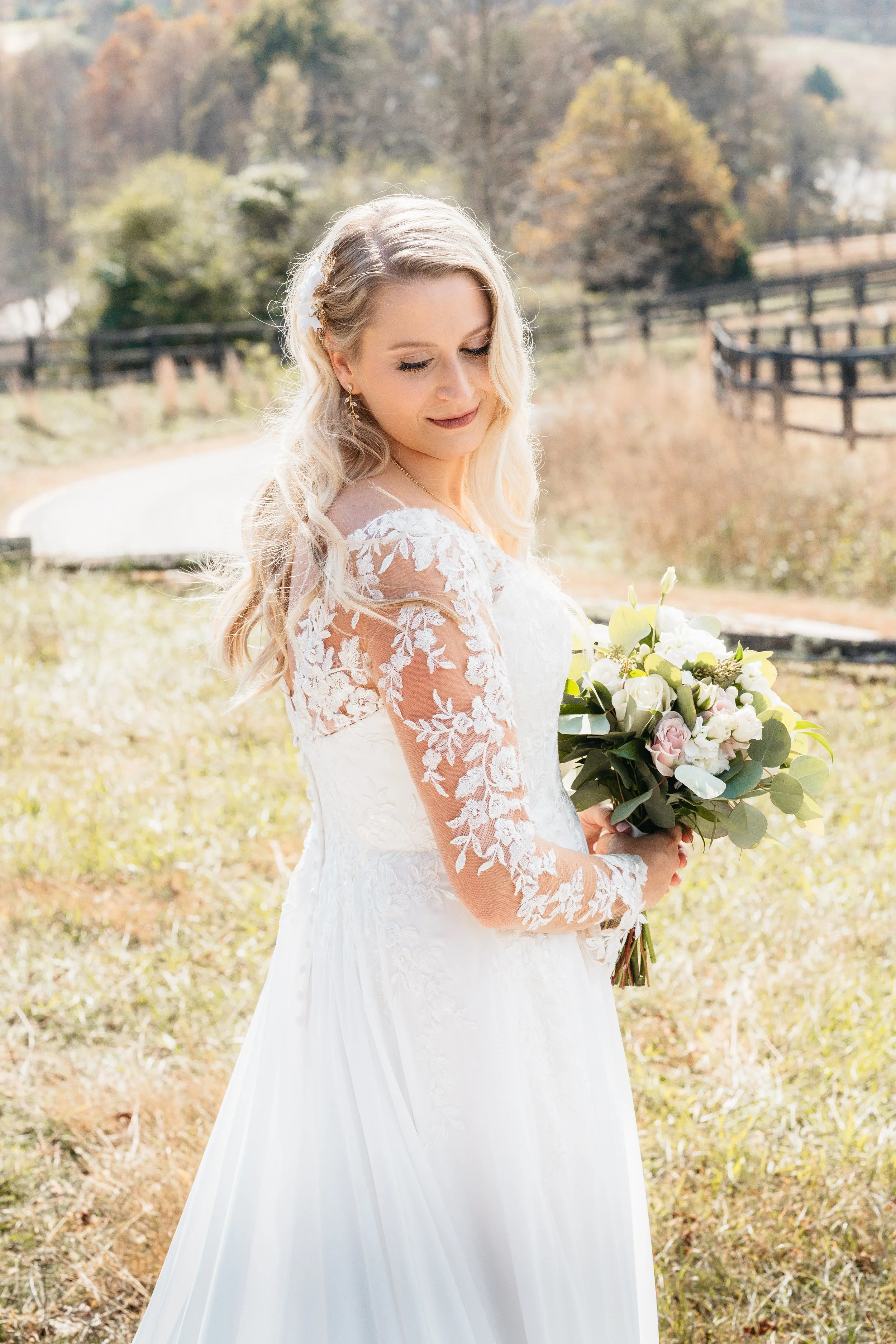 Georgia Mountain wedding venue with bride in a white lace wedding dress holding a bouquet of flowers outdoors on a sunny day with trees and a fence in the background.