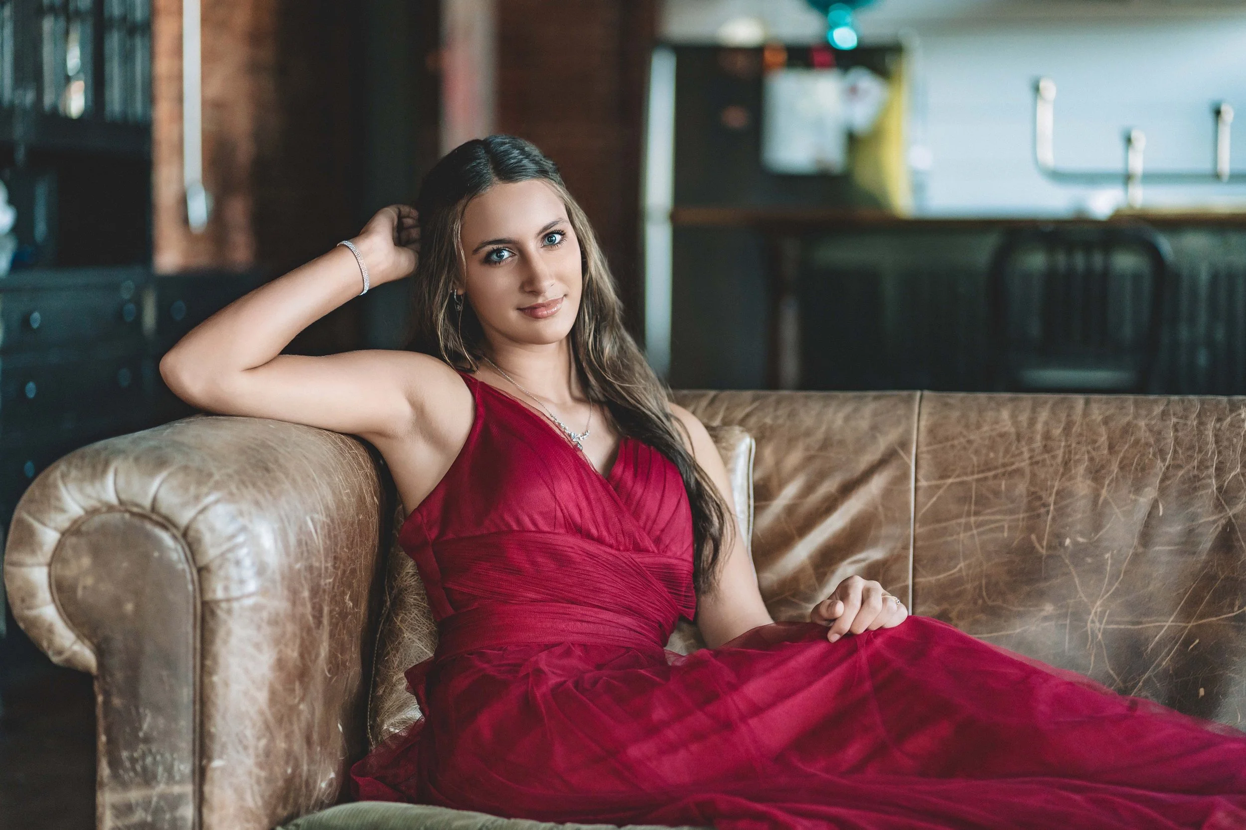 Young woman in a red dress sitting on a leather sofa in a cozy, rustic living room with dark furniture and a brick wall.