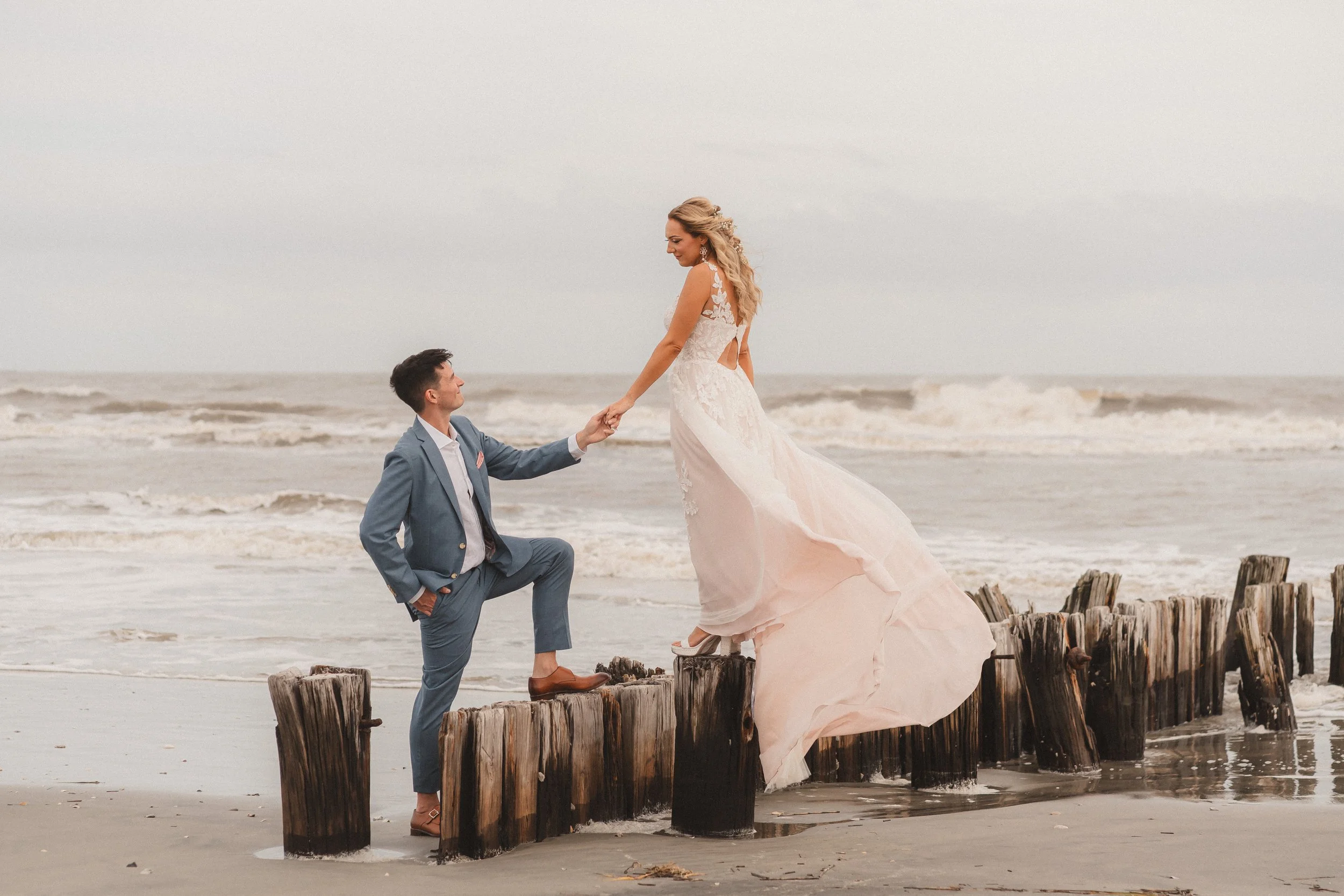 A couple dressed in wedding attire on a beach, with the groom kneeling and the bride standing on wooden posts, holding hands near the ocean.
