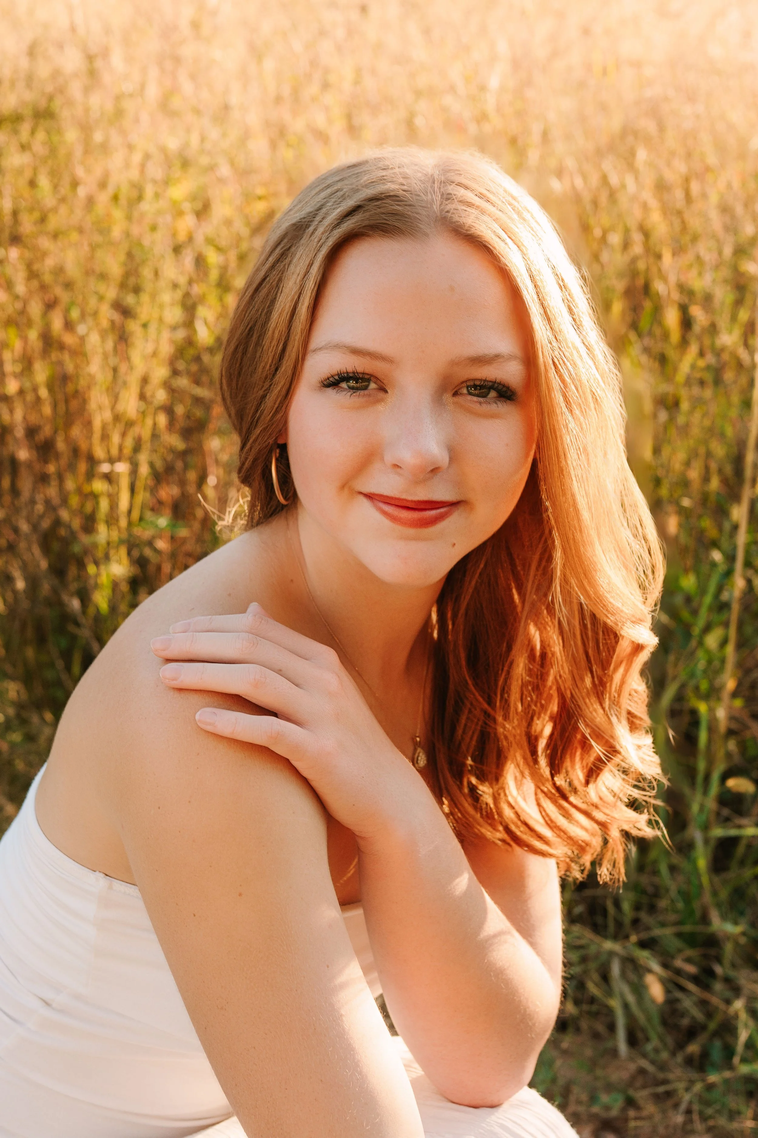 A young woman with long red hair and green eyes smiling at the camera, outdoors in a field of tall grass during sunset. Dreamy and elegant close up photo