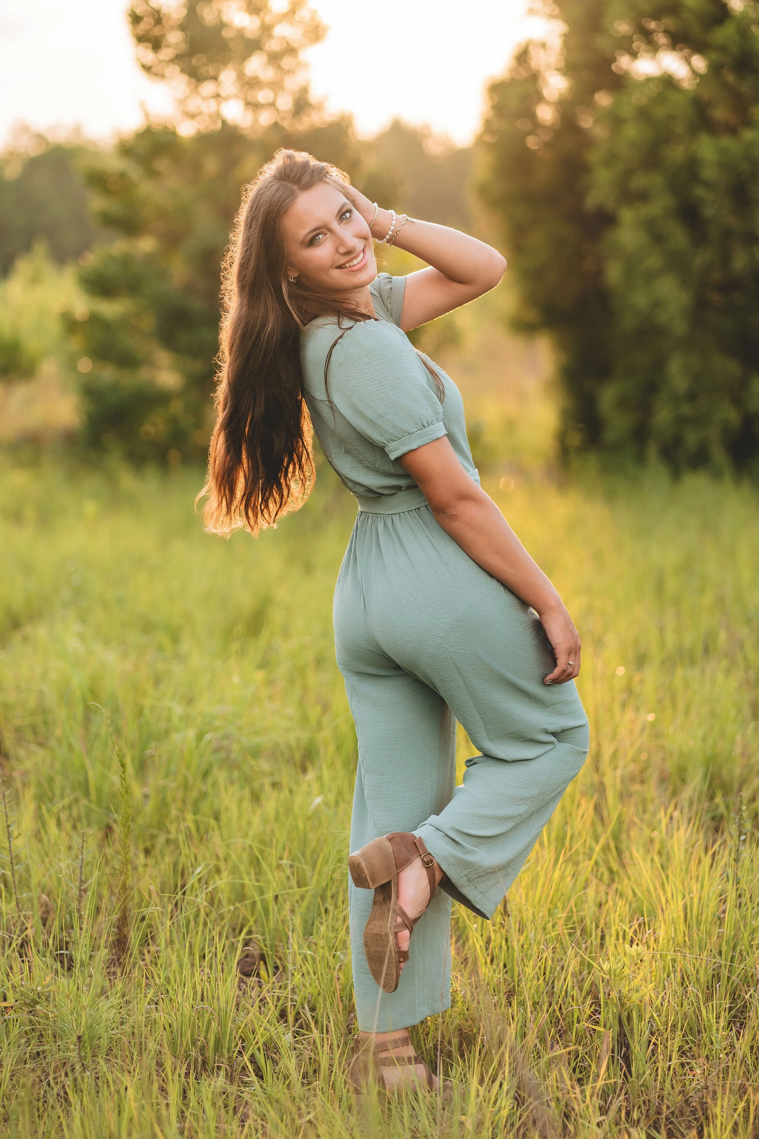 A woman with long brown hair posing in a green outfit and tan heels in a grassy field during sunset.