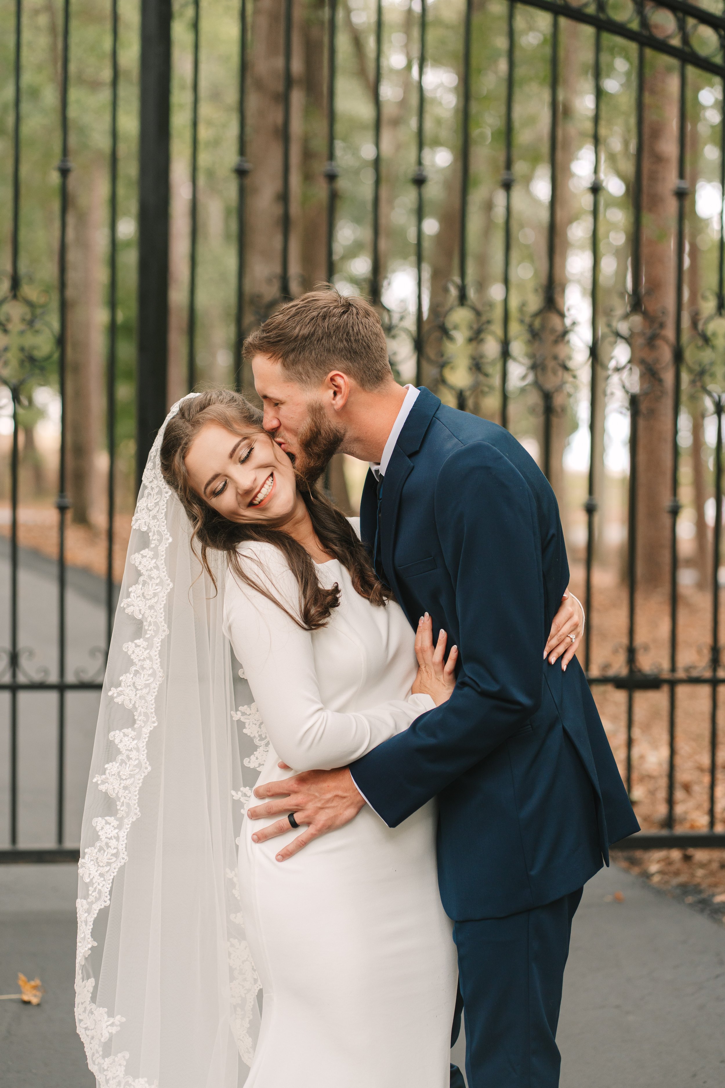 A bride and groom sharing a joyful moment outdoors, with the groom kissing the bride's forehead as she smiles happily. The bride is wearing a white wedding dress and a veil, while the groom is dressed in a navy blue suit.