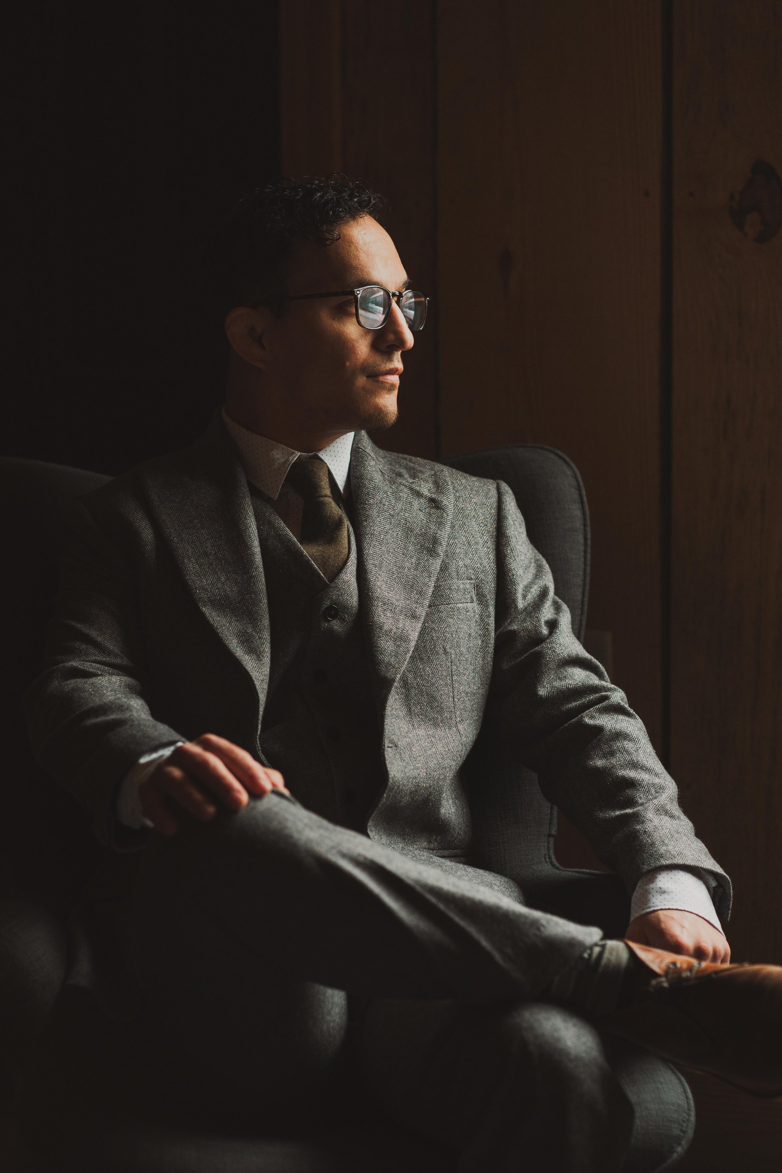 Moody portrait of Groom in a gray suit, glasses, and a tie sitting in a dark room with wooden walls, looking pensively out of the window.