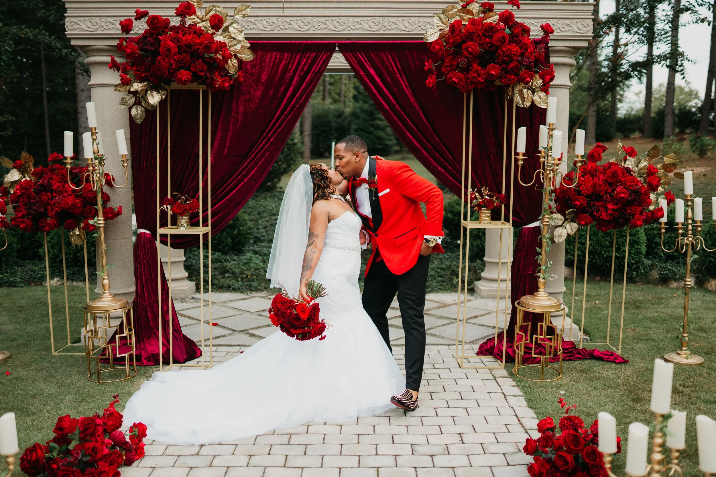 Valentine's day wedding decor, A bride and groom share a kiss during their wedding ceremony outdoors under a decorated arch with red roses and candles.