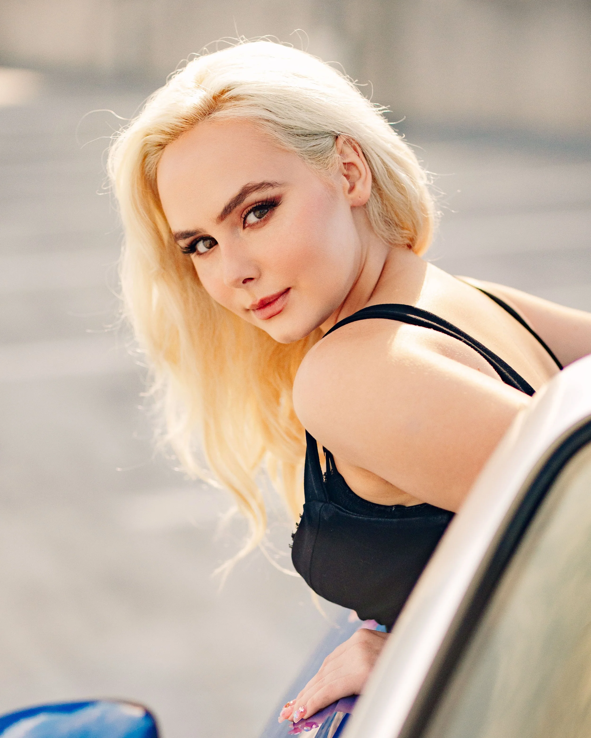 A woman with long blonde hair looking over her shoulder at the camera, wearing a black top, sitting on a white surface with a blurred outdoor background.