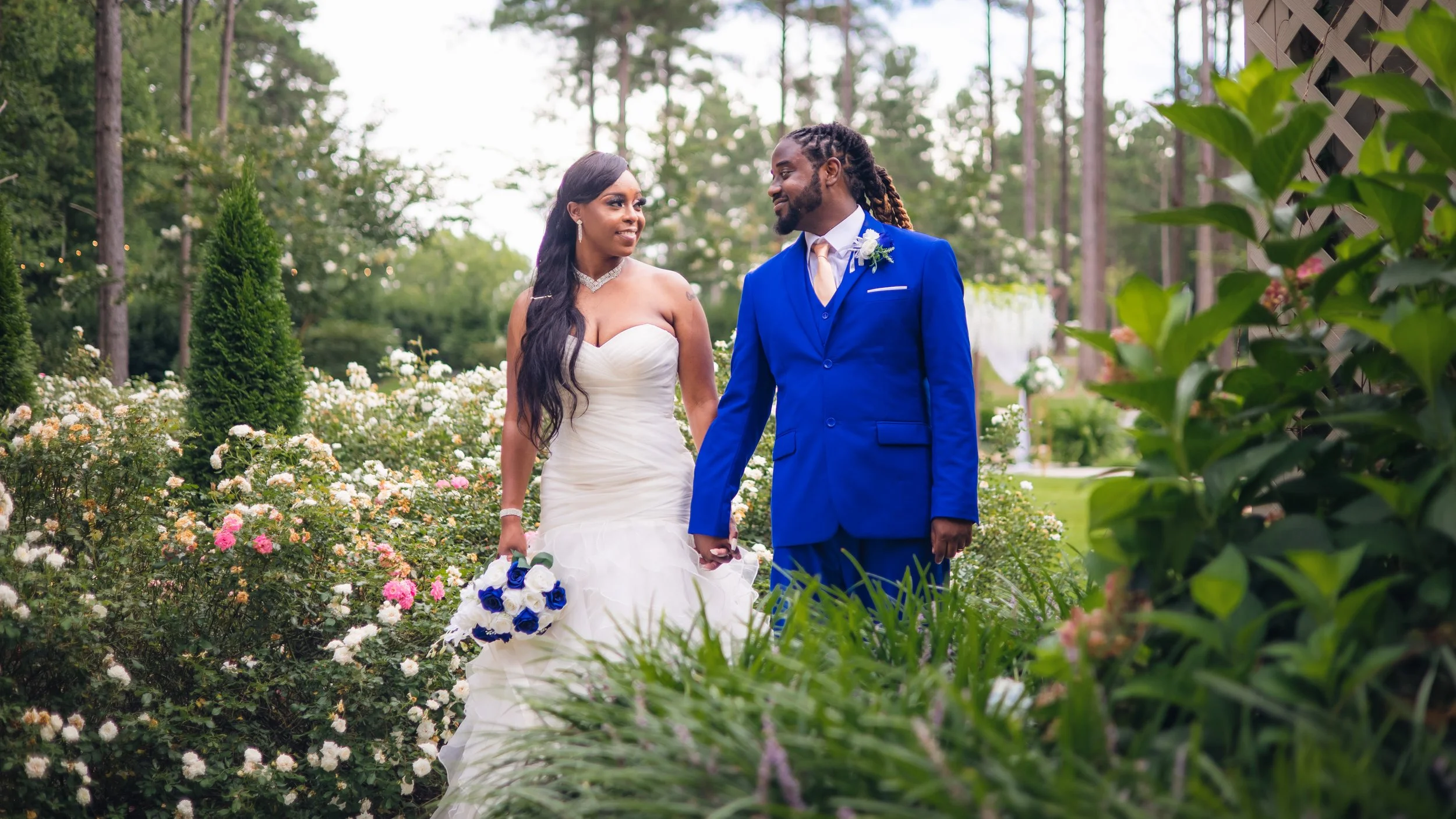 A bride and groom holding hands and walking through a garden with flowers and trees, with the bride holding a bouquet of white and blue roses.