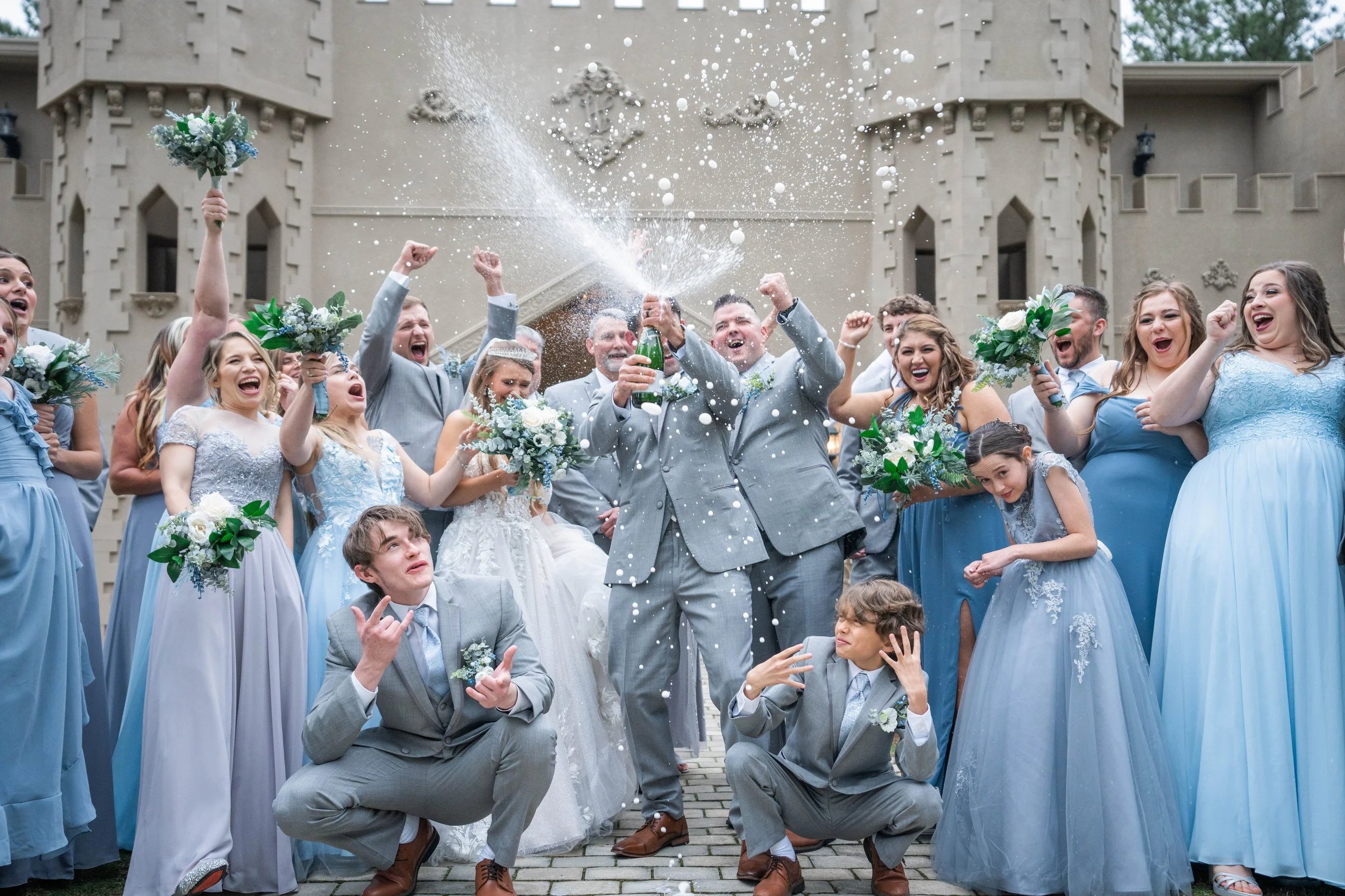 Wedding party celebrating with the bride and groom at the center as they splash champagne and cheer in front of Castle at Wildwood Gardens, fun champagne toast sendoff