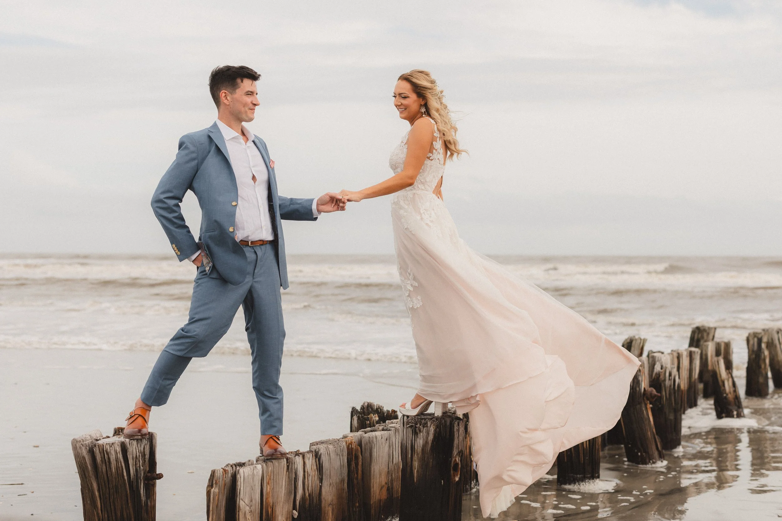 A bride and groom holding hands on a beach, standing on a wooden pier, with the ocean in the background.