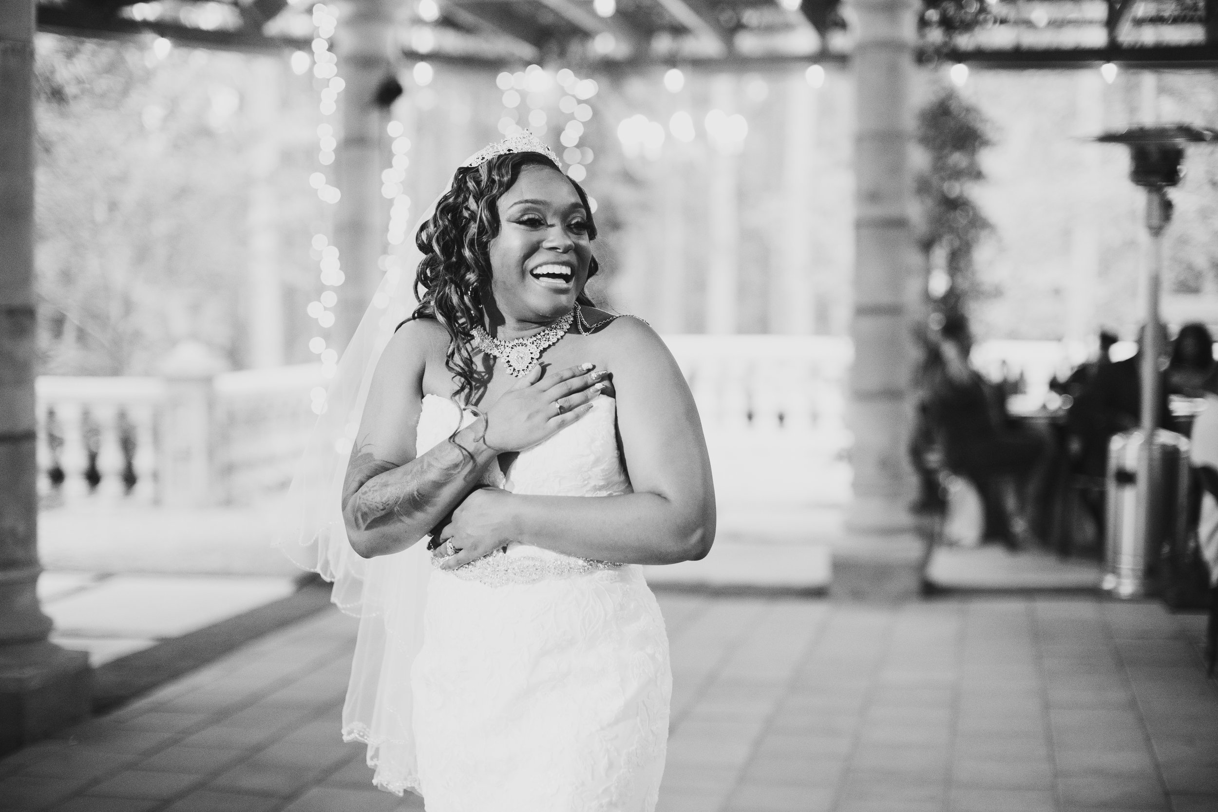 Emotional wedding day photo of a joyful bride in a wedding dress smiling and holding her chest with one hand, standing outdoors