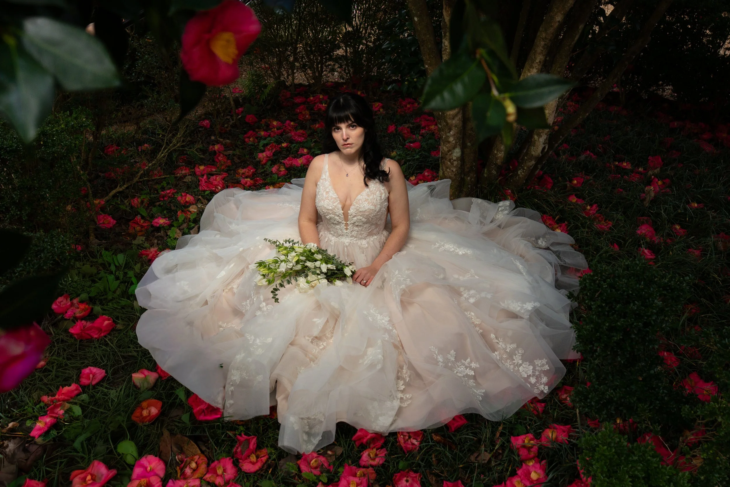 A woman in a wedding ballgown dress sitting on the ground surrounded by pink flowers and trees in a garden setting, vintage painting style photography
