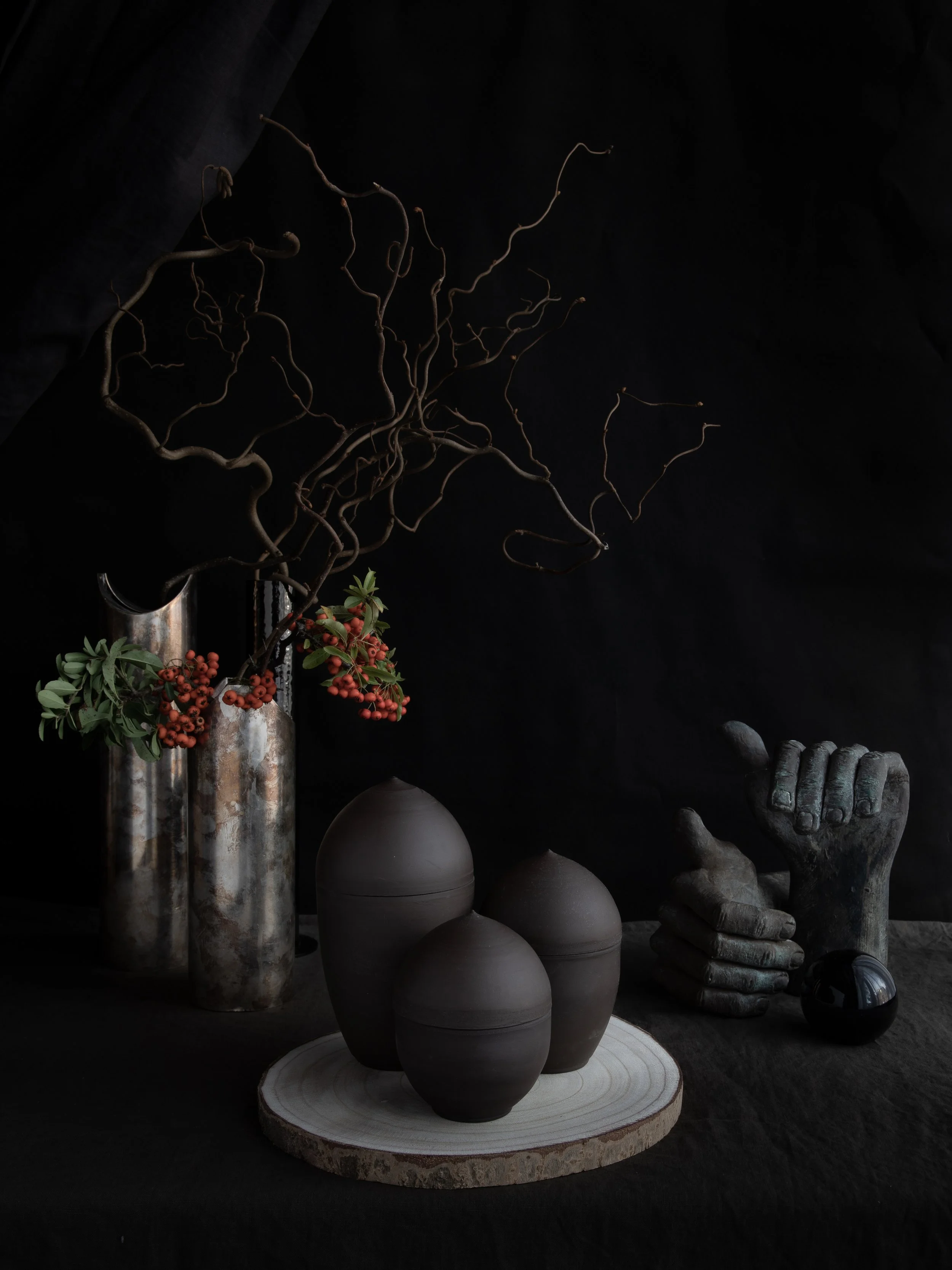 Black ceramic vases and a metallic hand sculpture with decorative branches and berries against a dark background.