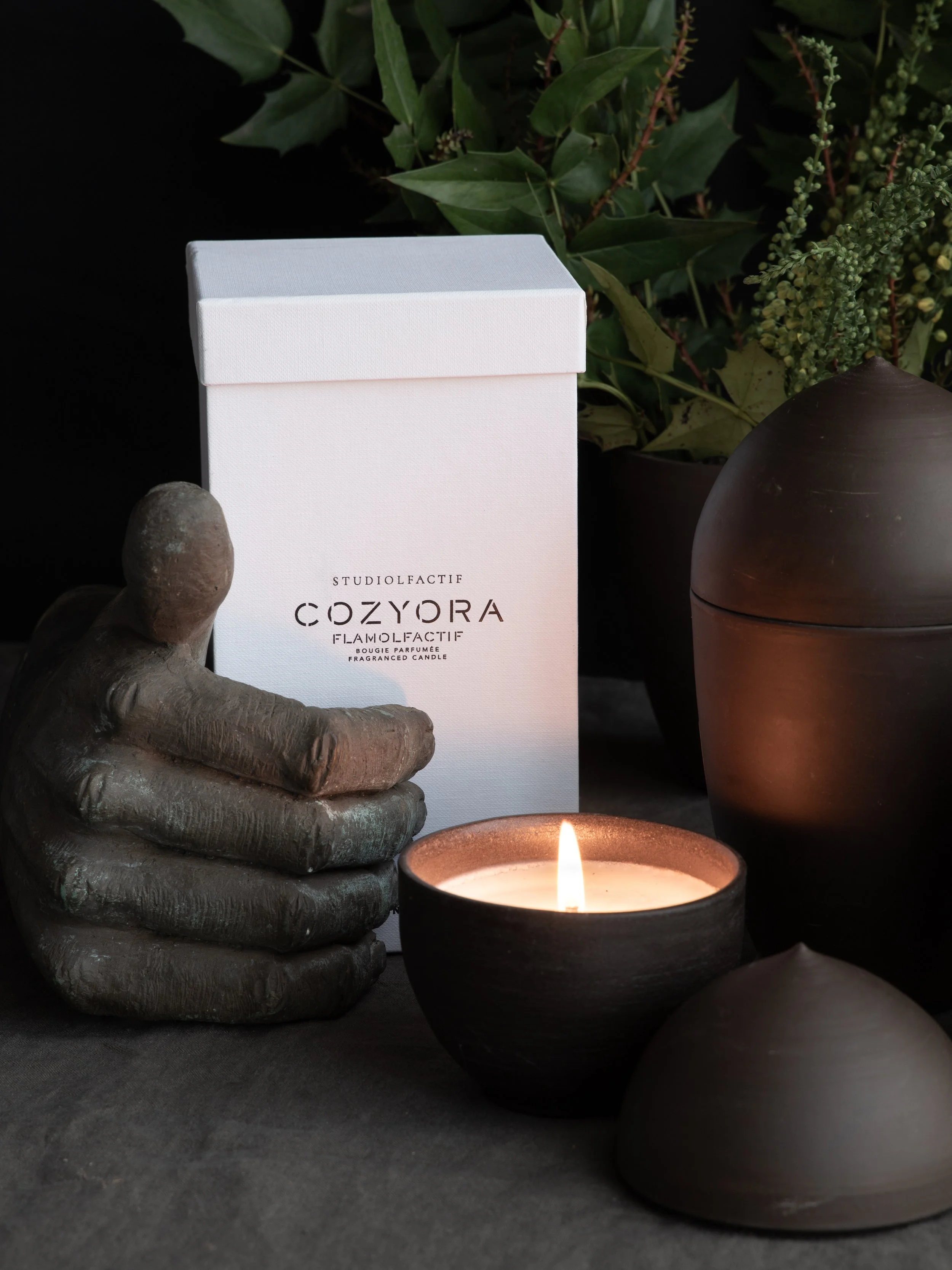 A white box labeled "StudioLfactif Cozyora" near a lit candle in a black holder, with a decorative hand sculpture and leafy greenery in the background.