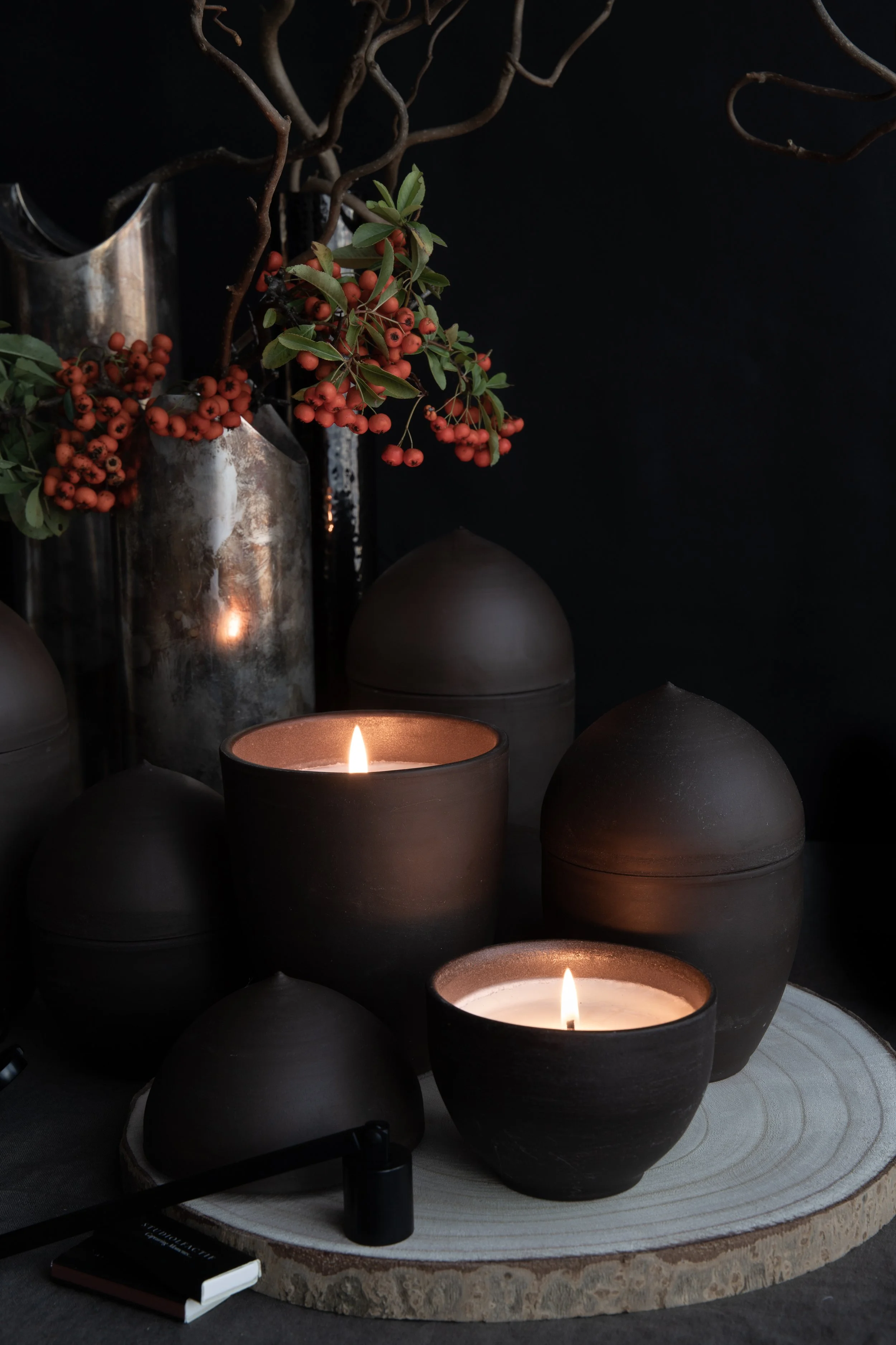 Lit candles in black containers on a wooden slice, with decorative branches and red berries in the background.