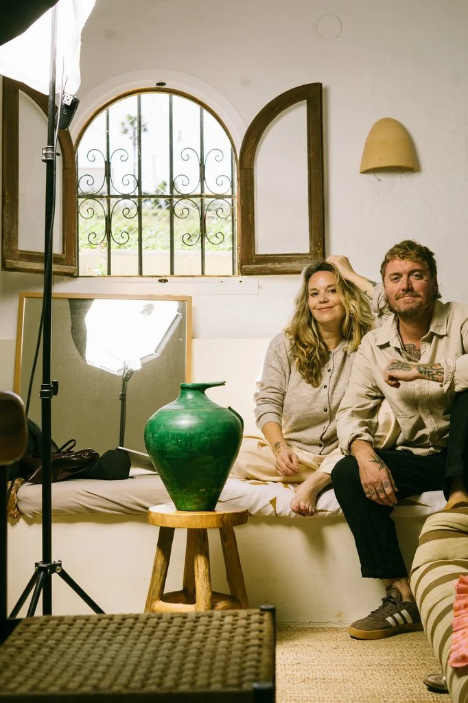 Two people sitting on a white couch in a room with a window, and a studio light setup in front of them. The woman has long blonde hair, holding part of her hair, and is smiling. The man has short hair, tattoos on his arms, and is sitting with a relaxed expression. There's a green vase on a small wooden stool in front of them, and an open window with wooden frames behind them.