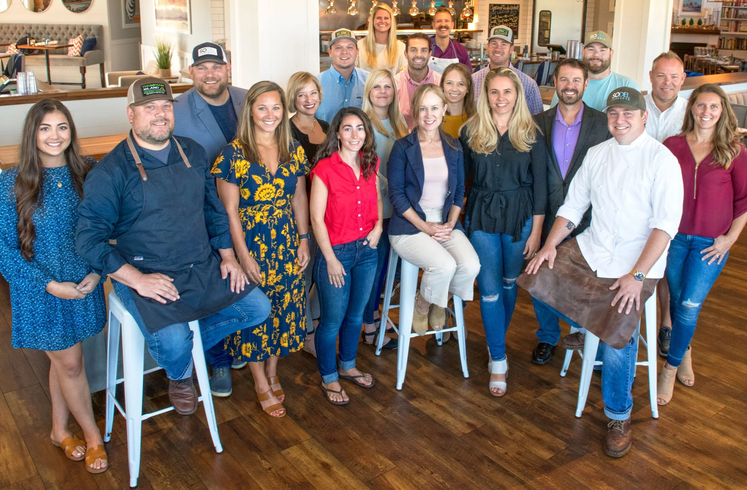 Group of smiling people in a restaurant with a bar and chalkboard menu in the background.