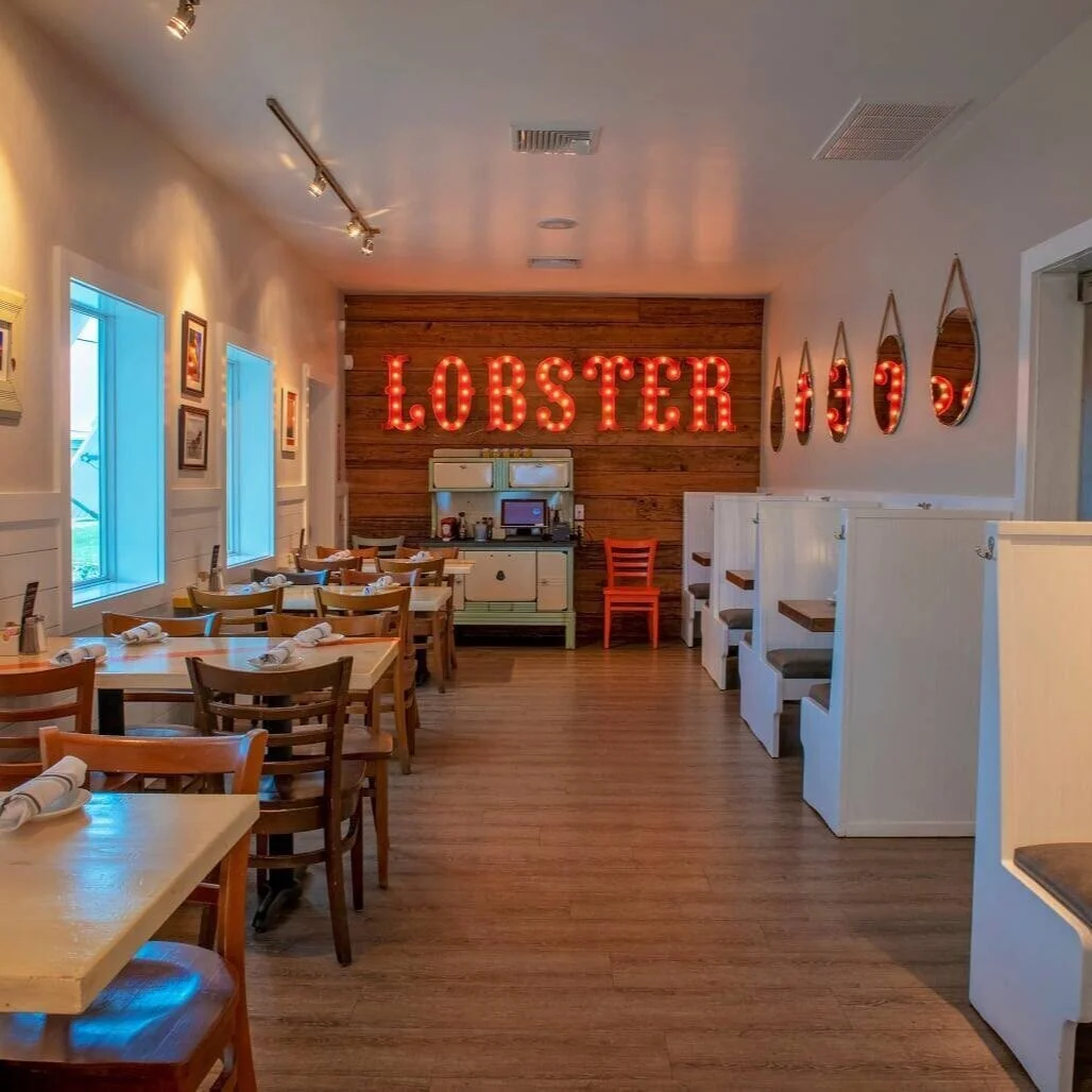 Empty restaurant with wooden floors and tables set with napkins and utensils, white booths along the right wall, decorative mirrors on the wall, three windows on the left, and a bright red neon sign spelling 'LOBSTER' on a wooden accent wall at the b