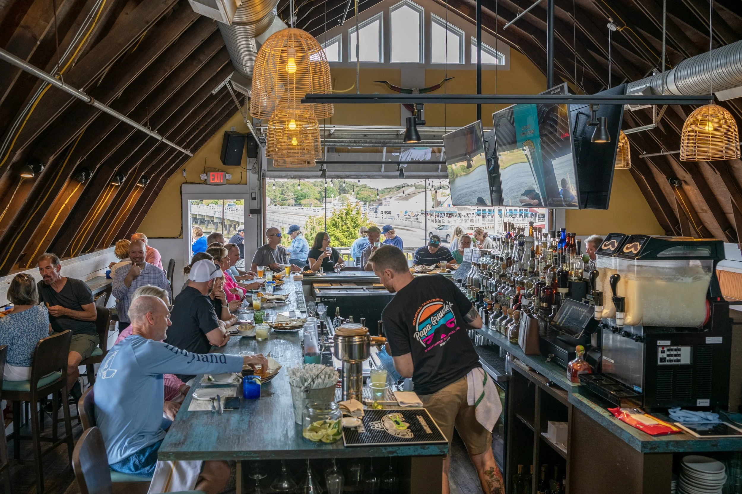 Inside a busy restaurant with people sitting at tables, a bar counter with liquor bottles, large windows, hanging wicker light fixtures, and ceiling ventilation pipes.
