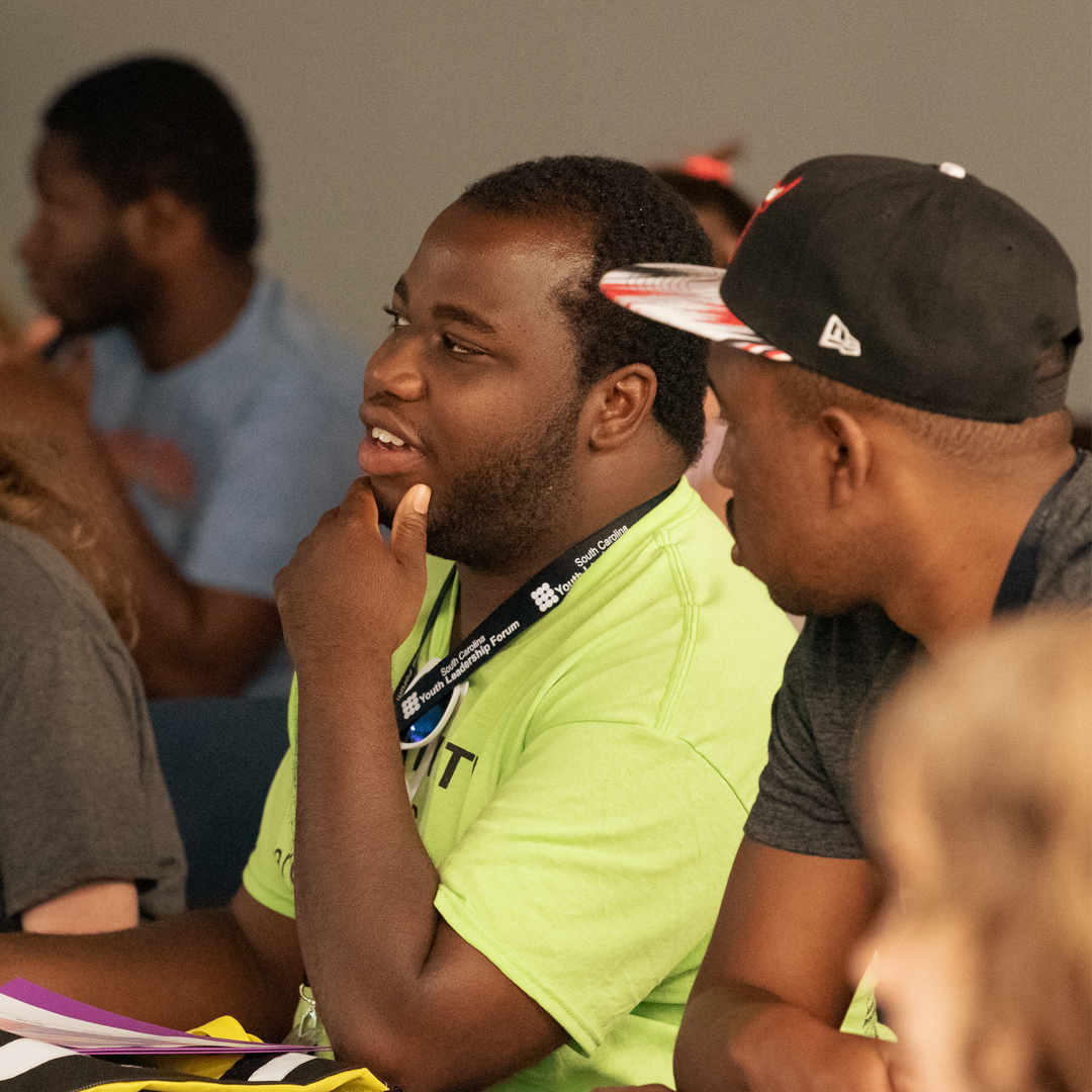 One 2019 delegate looks at the delegate sitting next to him, who has his hand resting on his chin as he looks to the front of the room.