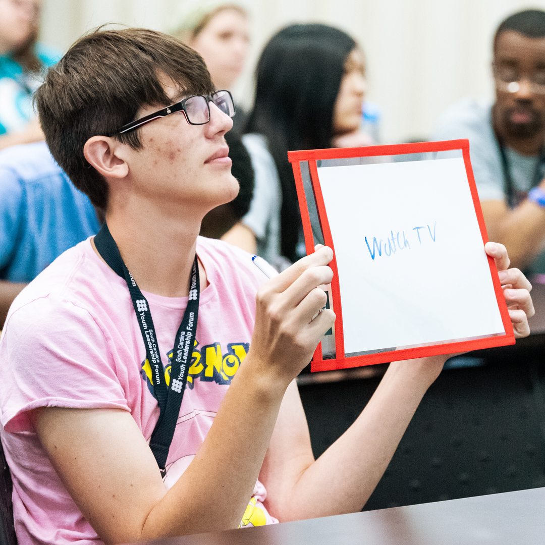 A delegate in a pink Pokemon shirt holds up a white board with the words “Watch TV” on it