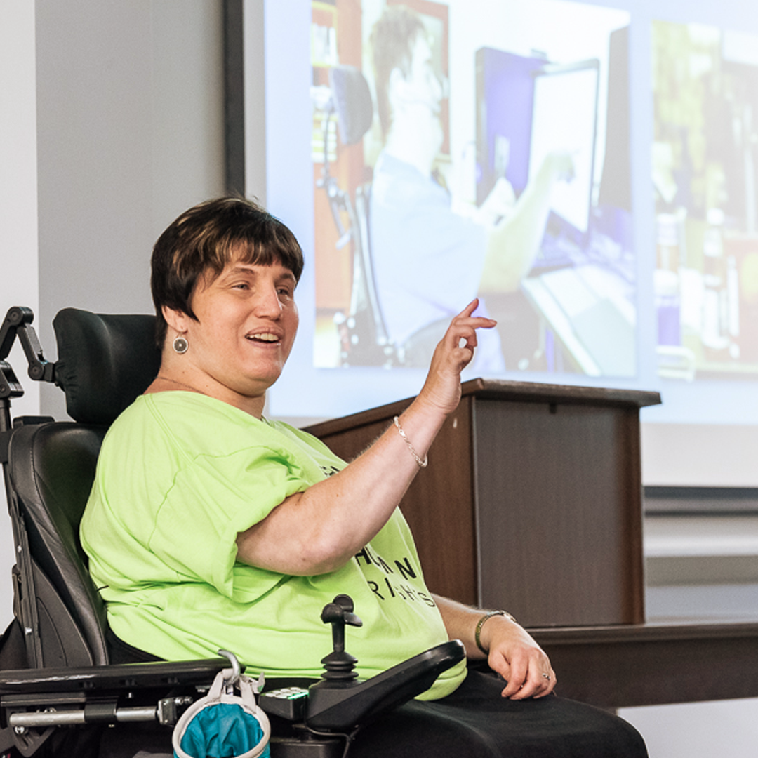 Staffer Dori sits in her wheelchair and speaks and points to her audience with a video playing on screen behind her.